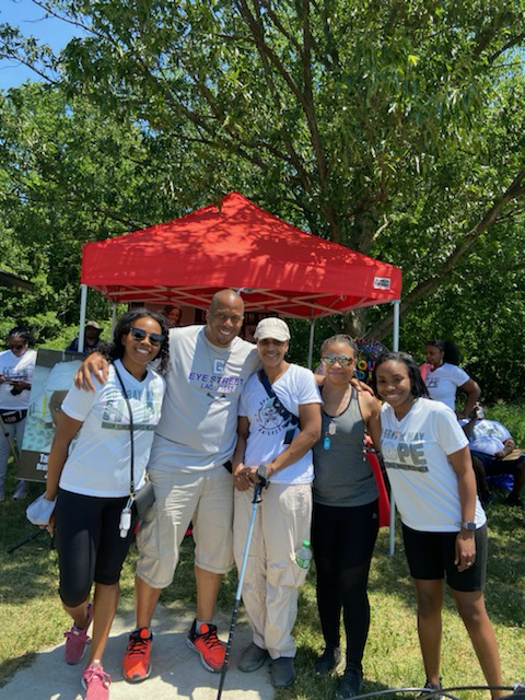 Group of five people standing outdoors in front of a red canopy tent, smiling at the camera, participating in a community event on a sunny day with green trees in the background.