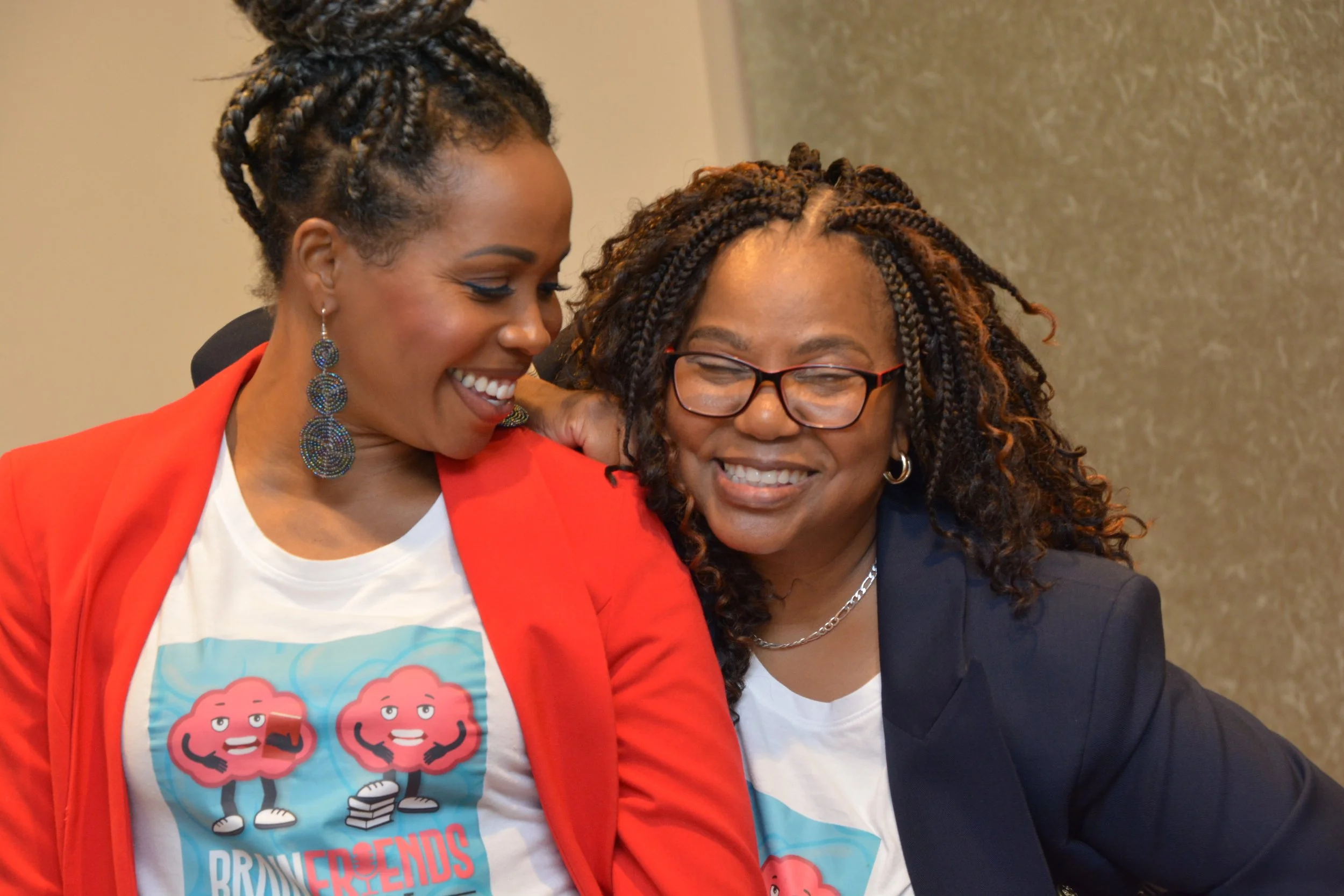 Two women smiling and talking to each other, dressed casually with one in a red blazer and the other in a dark blazer, both wearing T-shirts with a cartoon brain character and the text 'Brainiacs & Friends'.