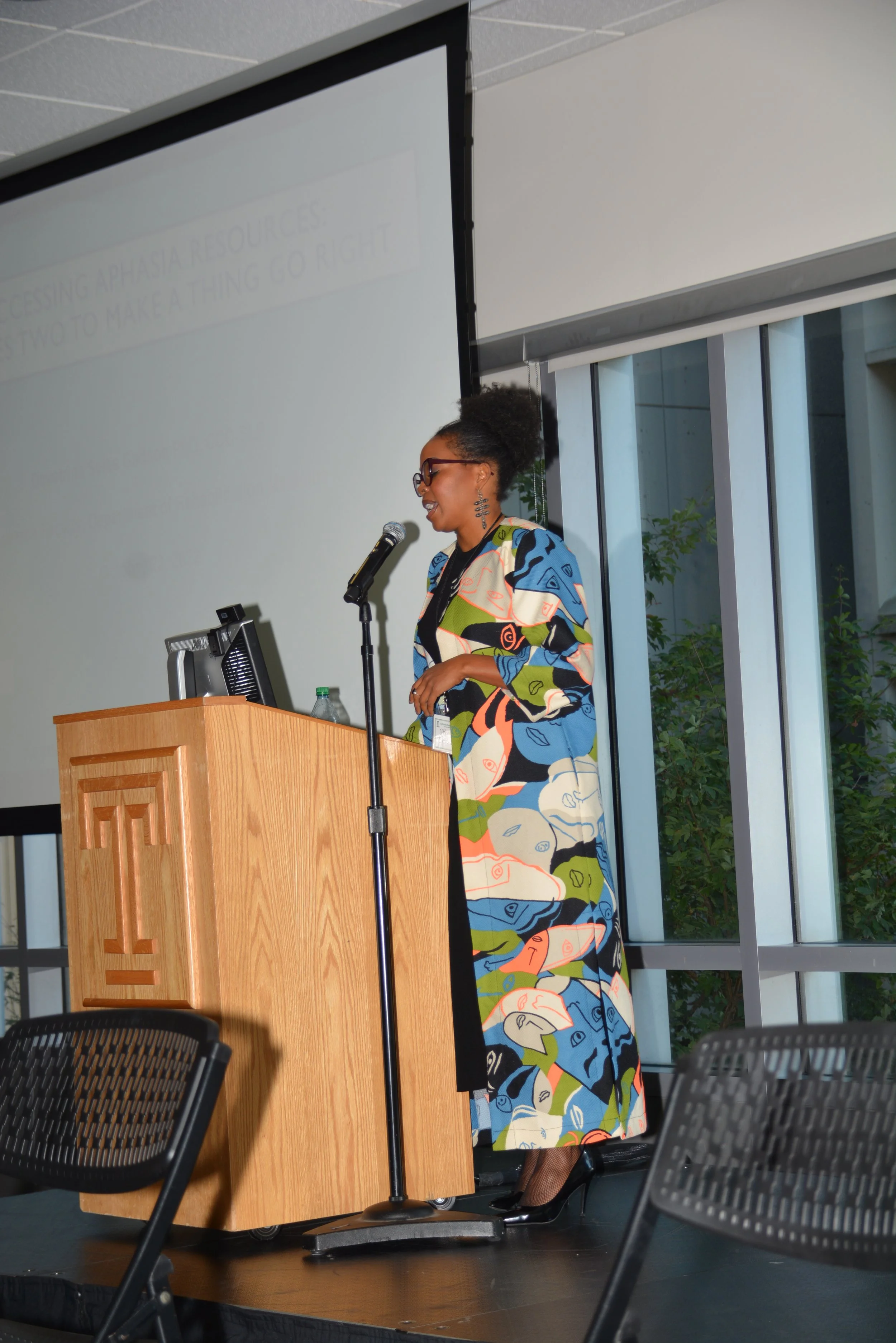 A woman in glasses and a colorful dress stands at a wooden podium, speaking into a microphone, with a large screen behind her displaying presentation slides.