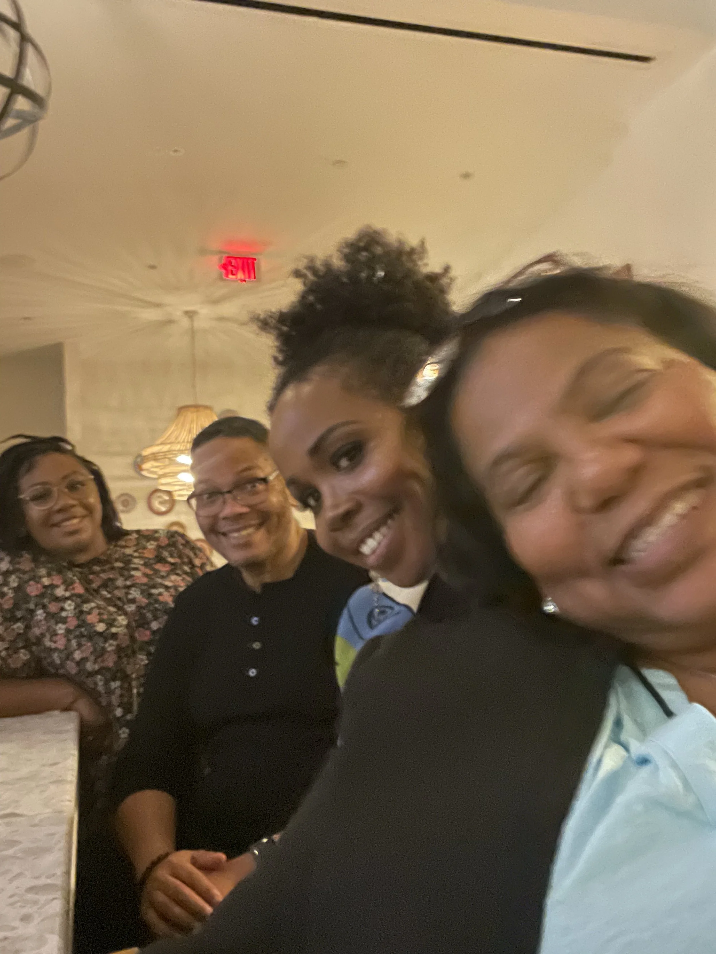 Four people smiling and posing for a photo in a warmly lit room, with a man wearing glasses and a black shirt, and three women with joyful expressions.