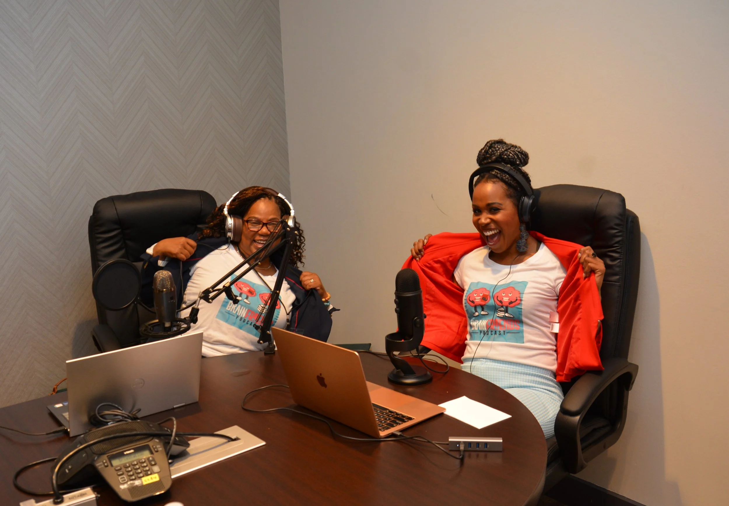 Two women sitting at a conference table, wearing headphones and laughing during a podcast recording. They have laptops, microphones, and recording equipment in front of them.