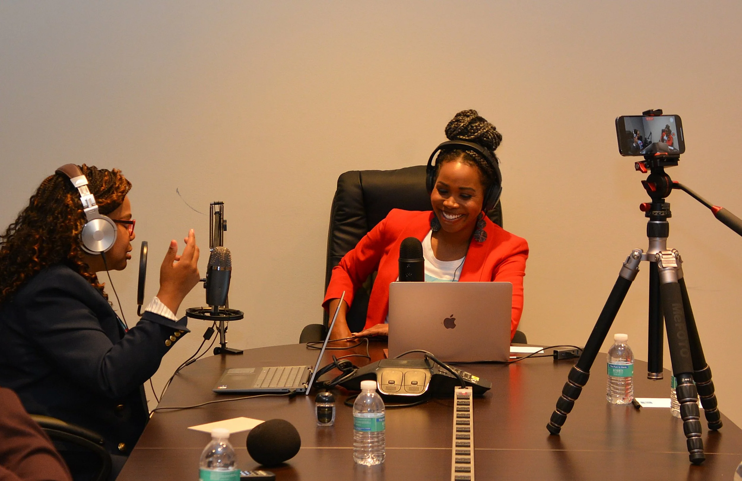 Two women conducting a podcast or interview in a studio with microphones, a camera on a tripod, laptops, and water bottles on the table.