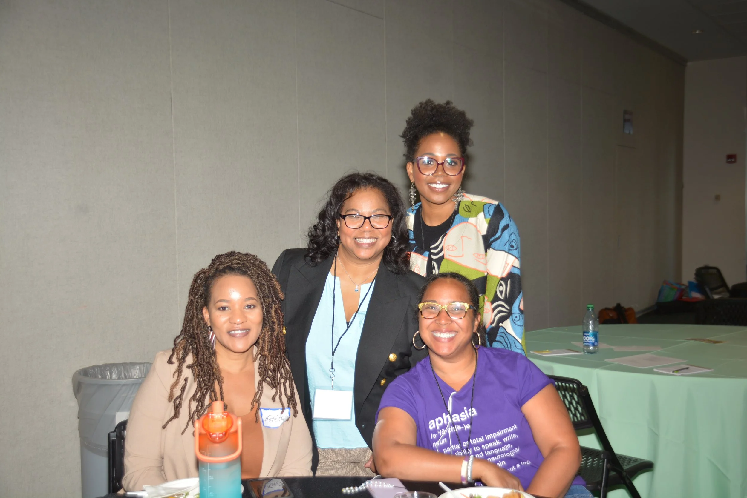 Four women at a conference or event, smiling and posing for a photo, seated at a table with water bottles and papers, in a room with a plain wall background.
