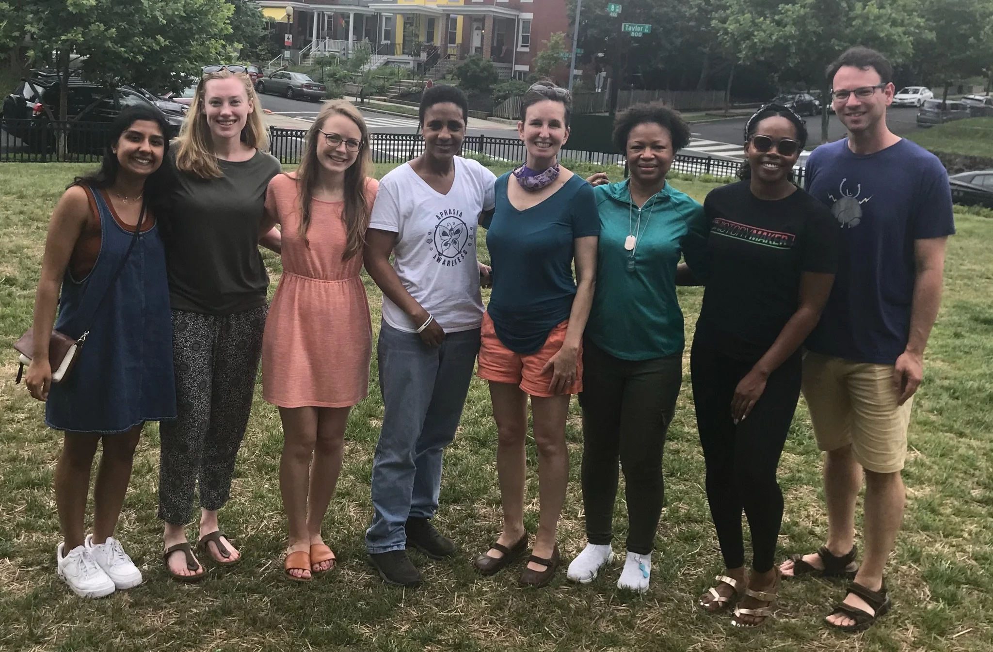 Group of nine diverse women and one man standing outdoors on grass, smiling at camera with a residential street and trees in the background.