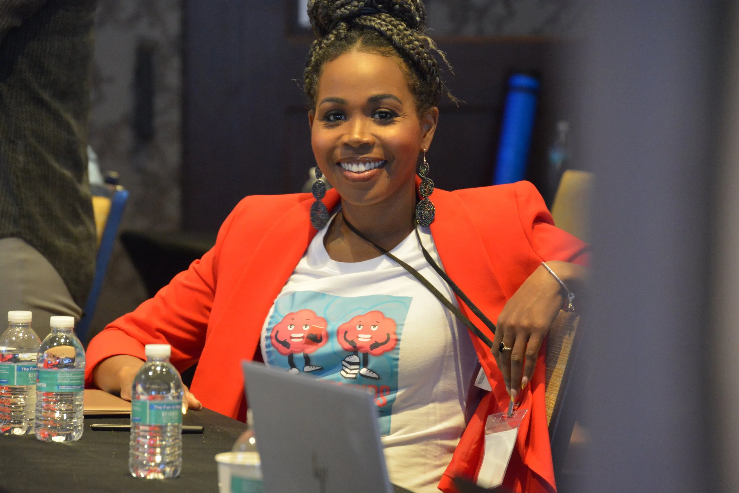 A woman with dark braided hair smiling, wearing a red blazer and earrings, sitting at a table with water bottles and a laptop.
