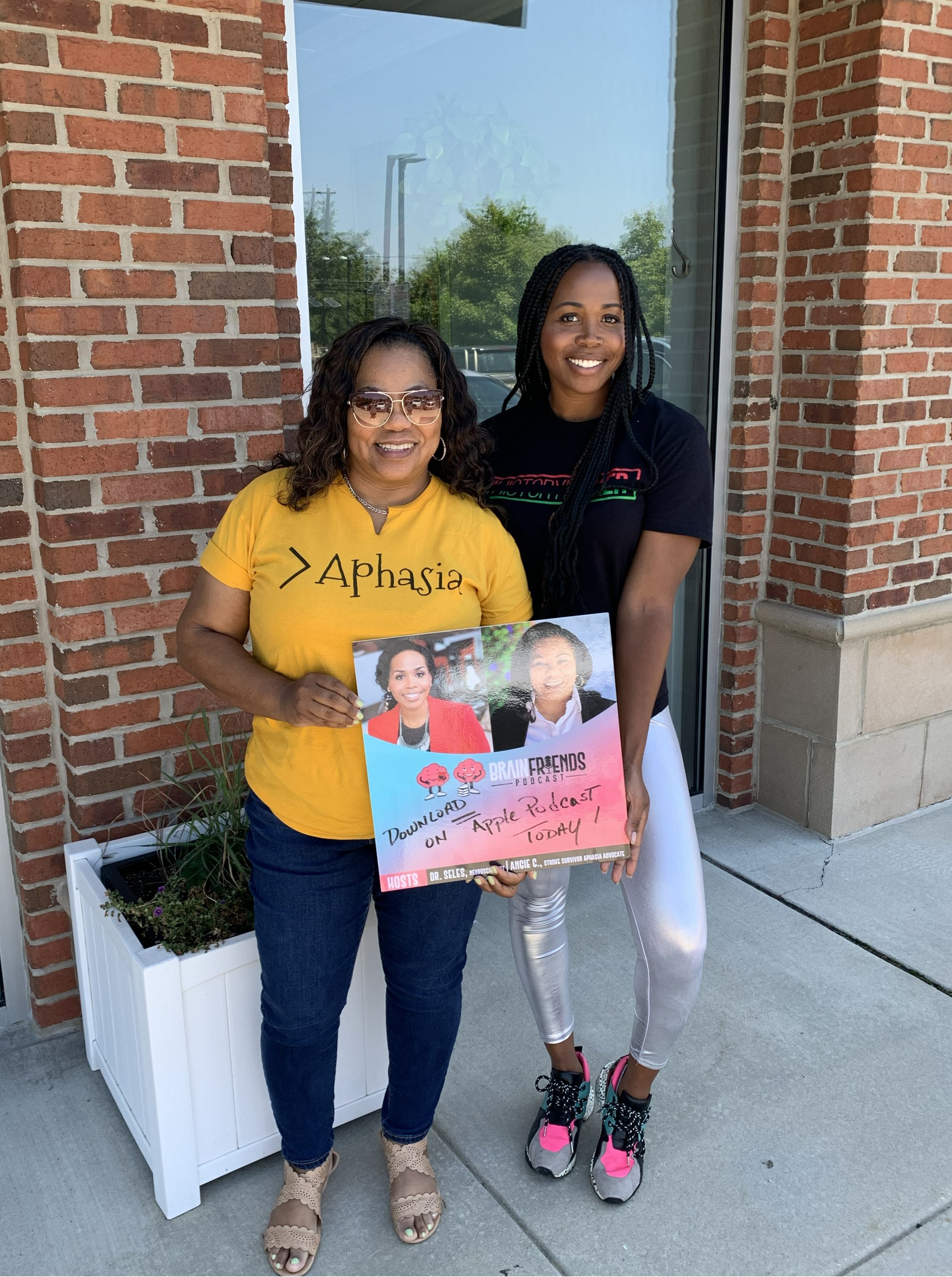 Two women standing with a sign in front of a brick building. The woman on the left is wearing a yellow shirt with the word 'AphasIa' and sunglasses, and the woman on the right is wearing a black t-shirt with colorful text and silver leggings. The sign features two photos of women and text about a podcast on Apple Podcasts, including the names Dr. Seles and Langie C.