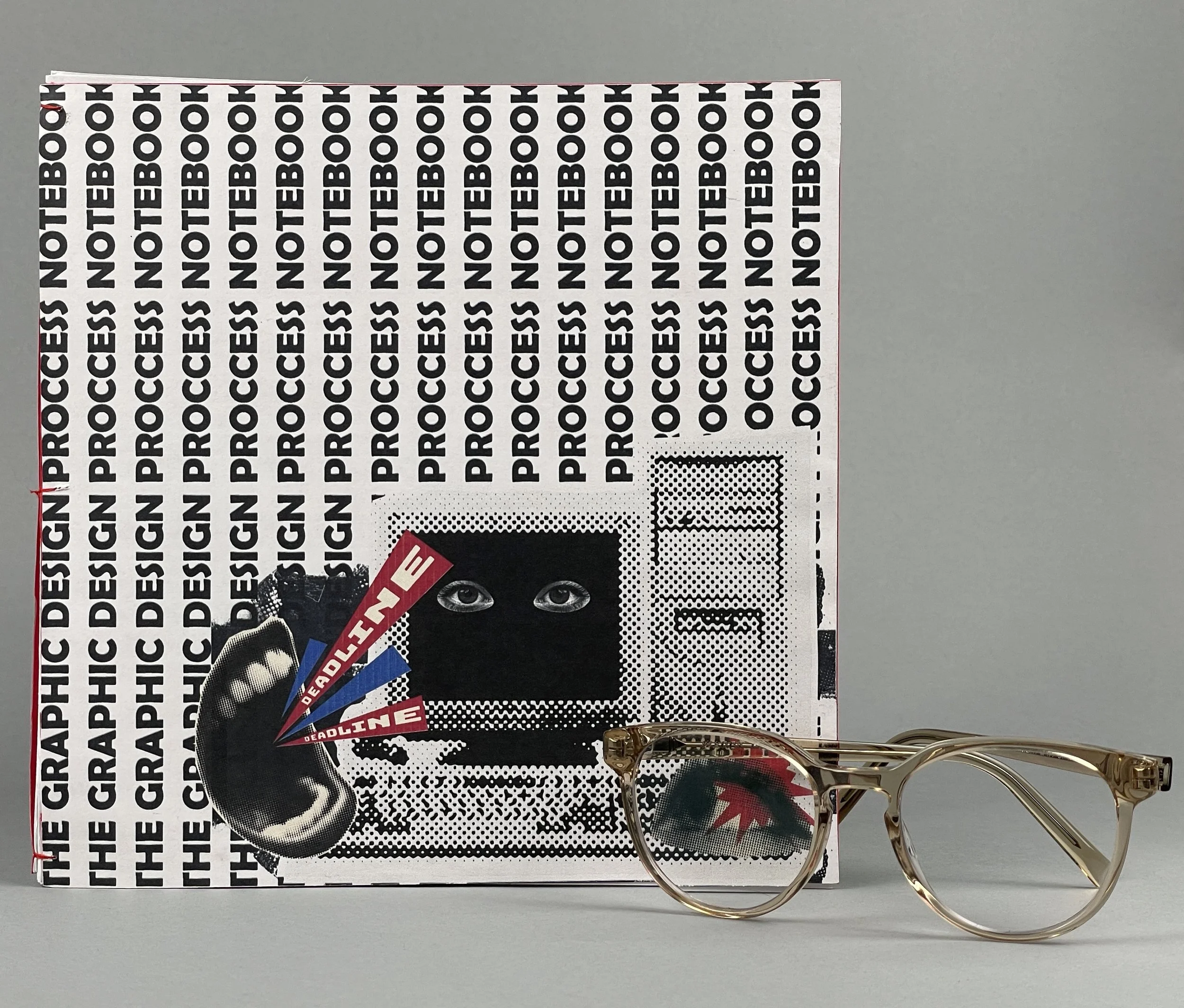 A graphic design book titled 'The Graphic Design Process' leaning against a gray background, with a sticker depicting a person with eyes, holding a pointed object labeled 'Deadline' and 'Deadlines'. A pair of clear-framed eyeglasses with a leaf patte