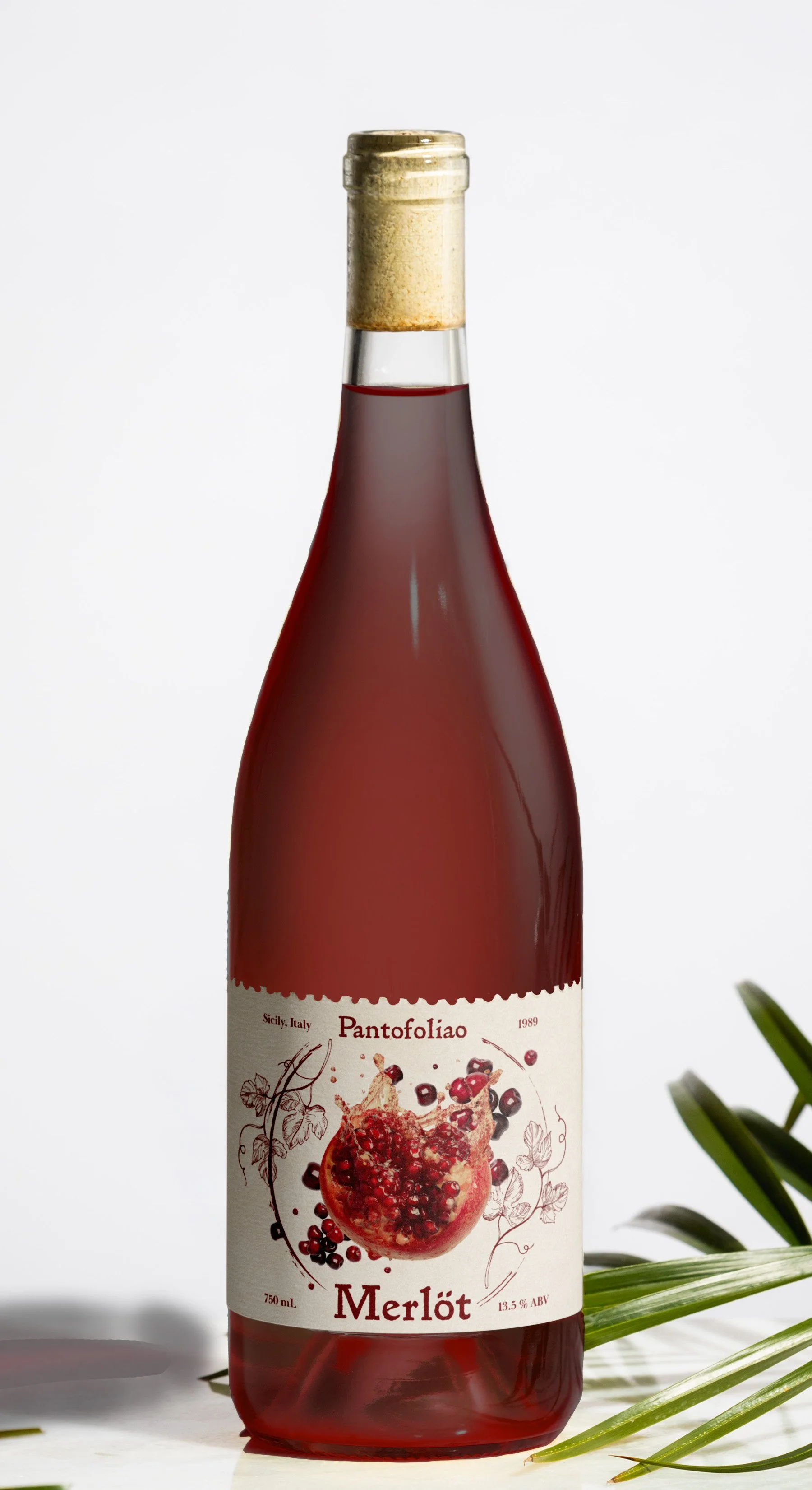 A bottle of Merlot wine with a label featuring an image of pomegranate seeds and berries, placed on a white surface with some green palm leaves nearby.
