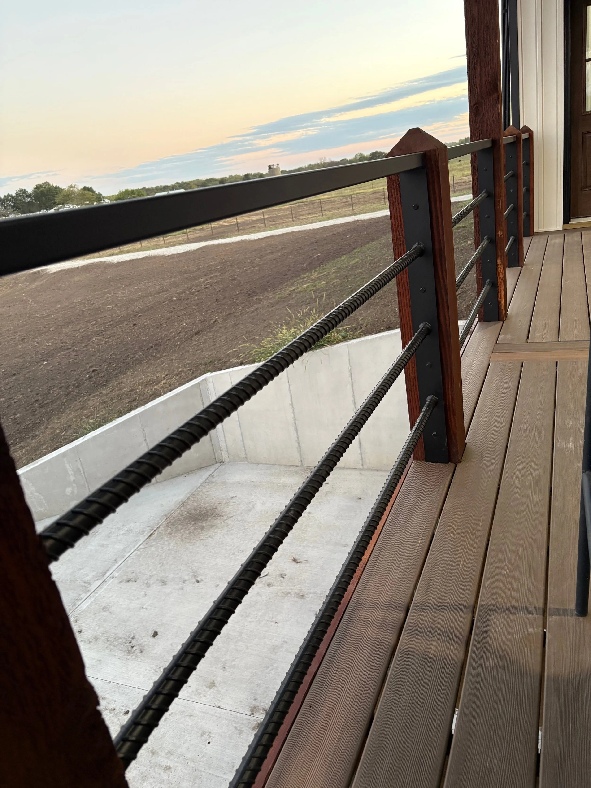 Wooden porch railing overlooking empty farmland with trees and a cloudy sky at sunset.