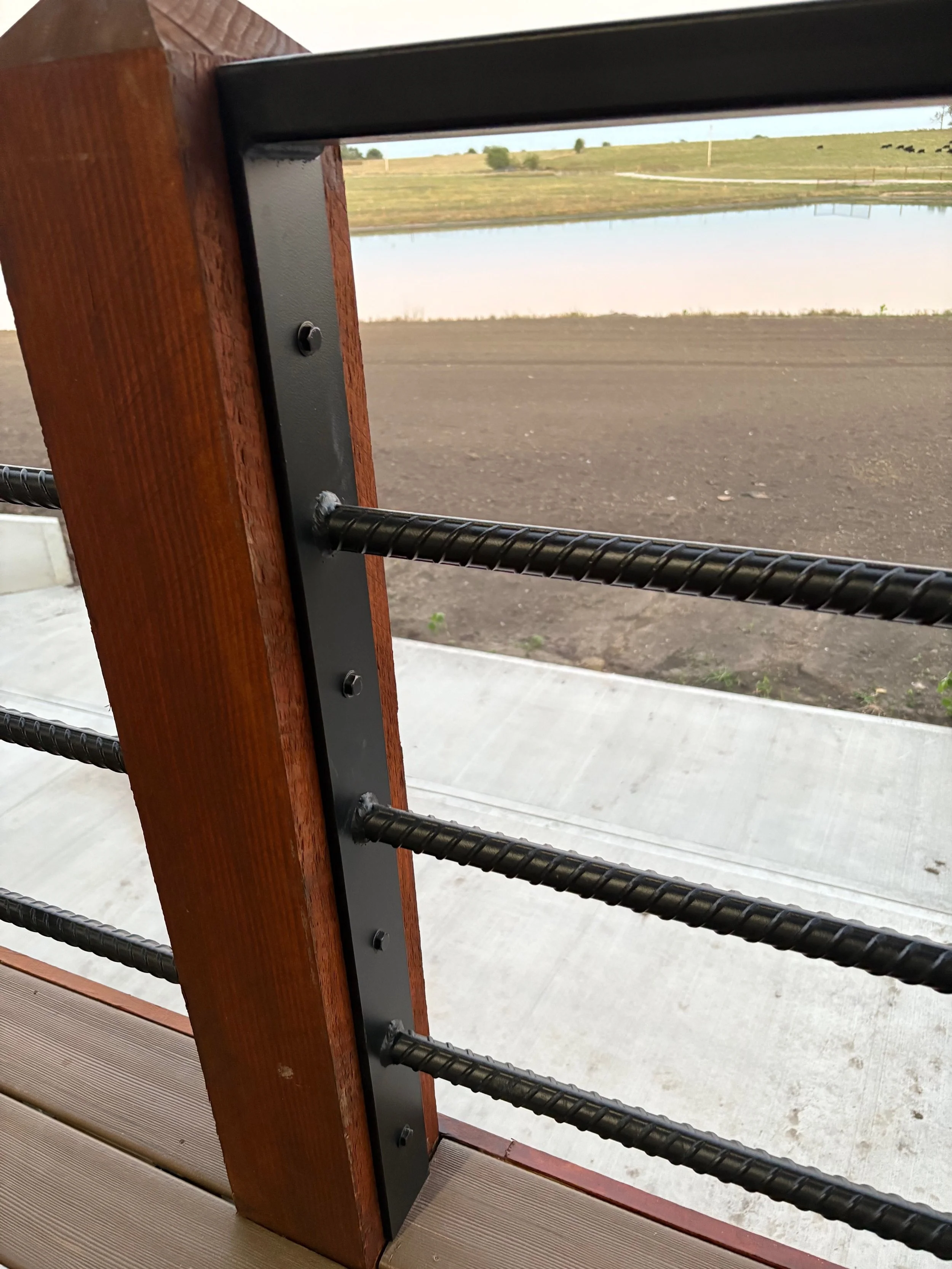 Close-up of a wooden railing with black metal bars and posts overlooking a dirt path, a small lake, and green open field with cows in the distance.