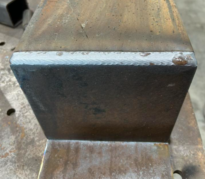 Close-up of a metal piece with a welded seam and a small rust spot, situated on a workbench or industrial surface.