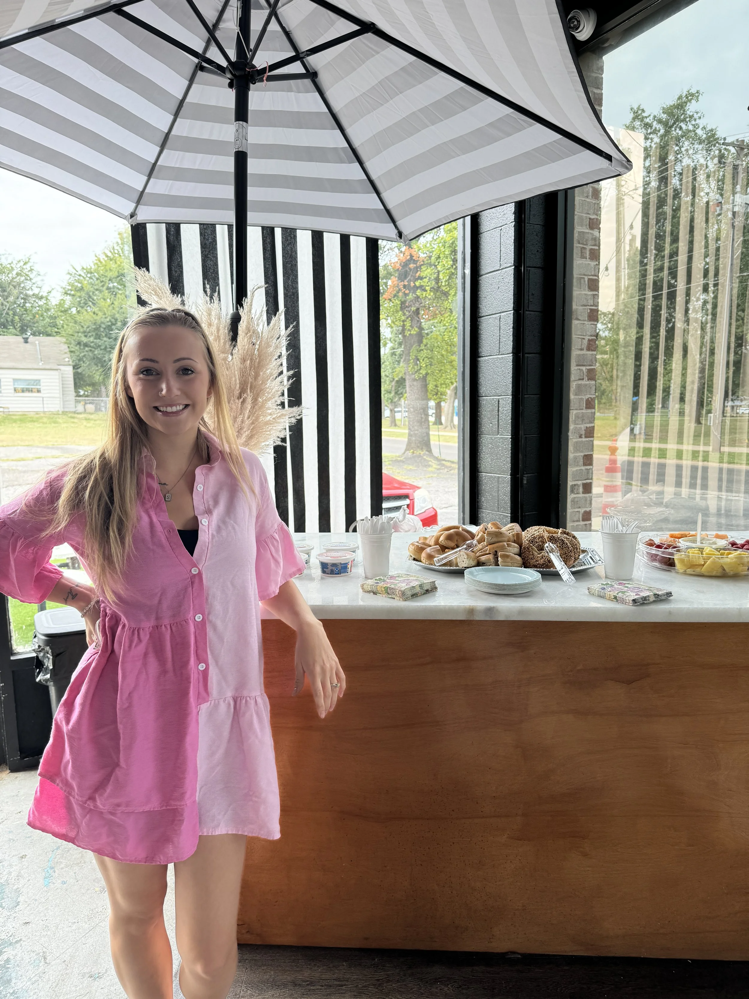 Smiling woman in pink dress standing at a table with assorted donuts, fruit, and condiments, inside a cafe with black brick walls and large glass windows.