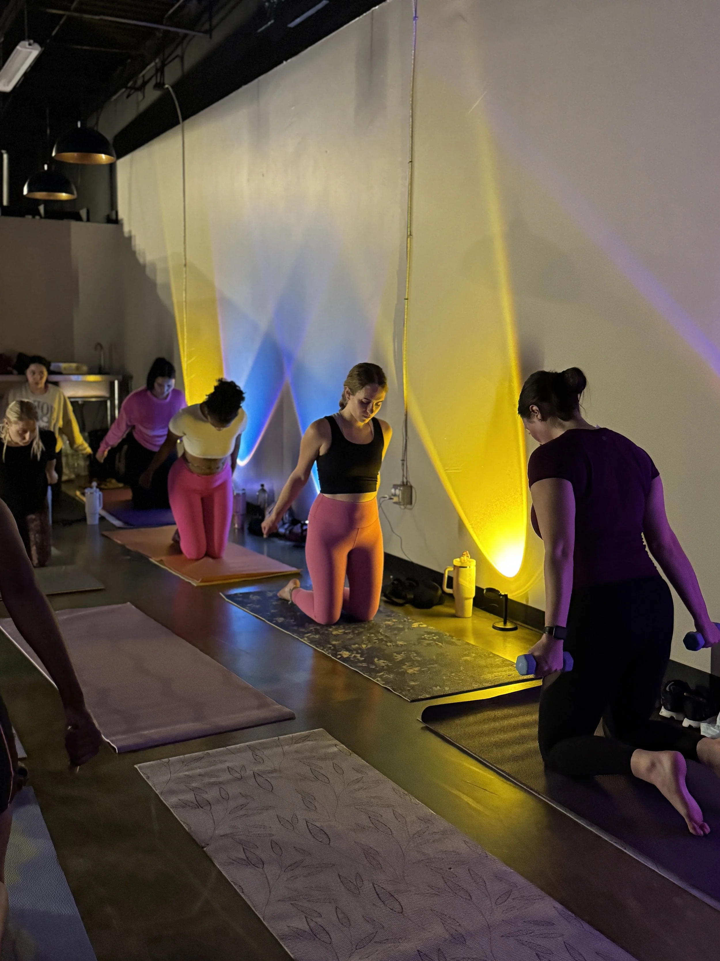 People participating in a yoga class in a dimly lit studio with colorful lighting, some kneeling on yoga mats and a woman holding small dumbbells.