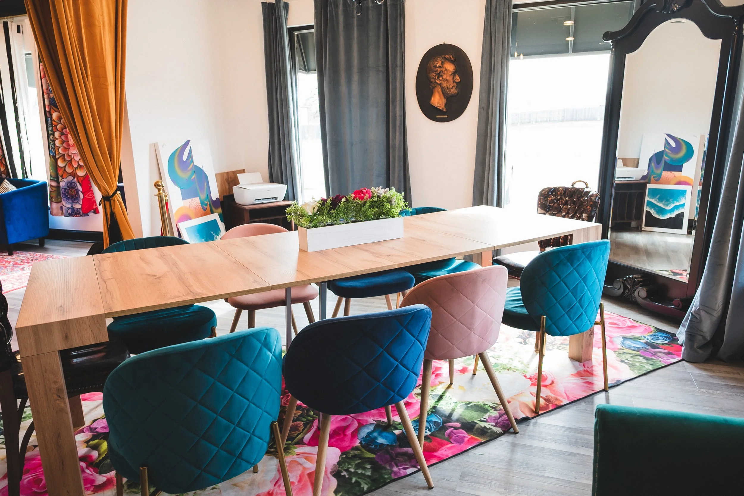 A dining room with colorful chairs around a wooden table, floral rug, and artwork.