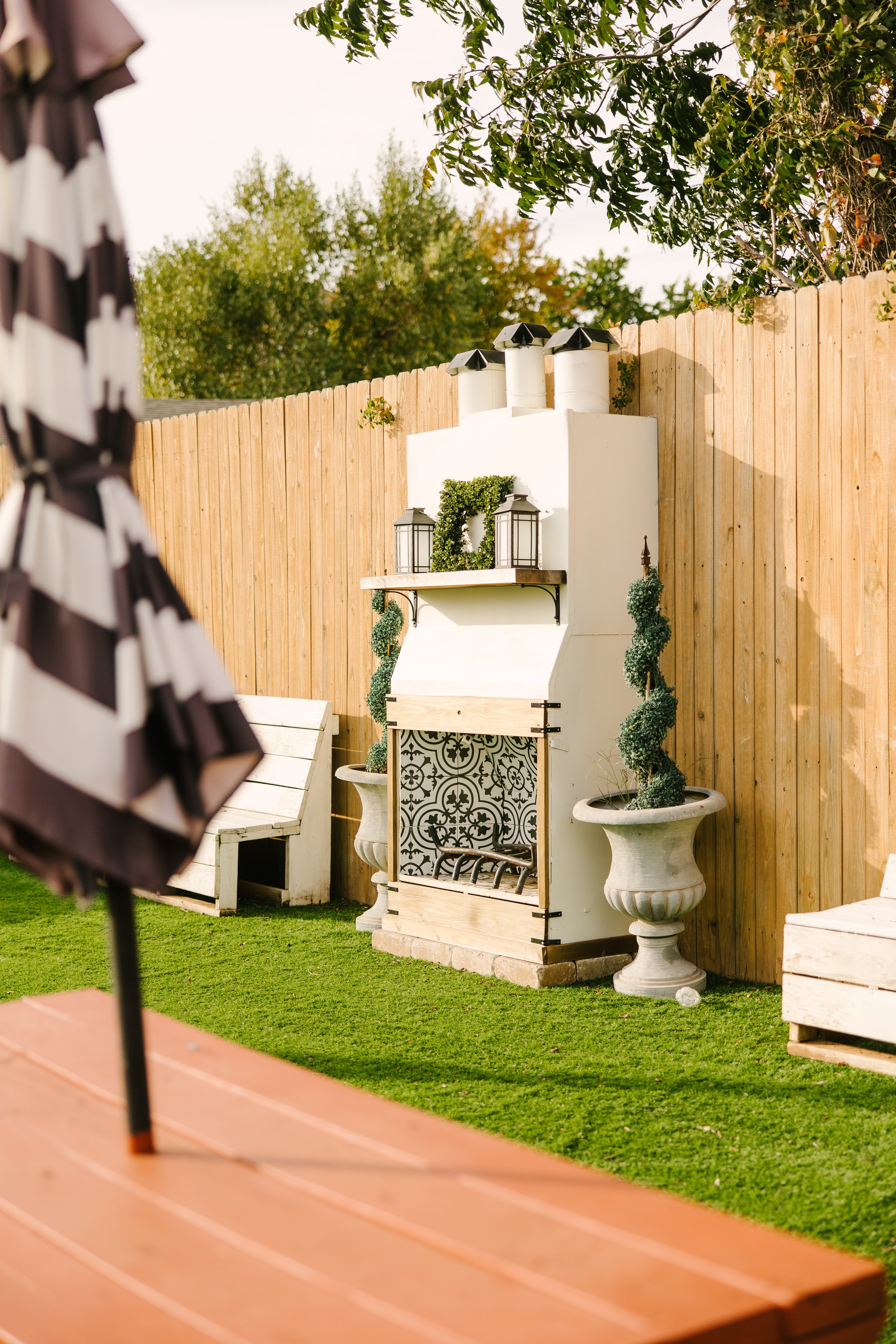 A backyard patio with a decorative white outdoor fireplace flanked by potted topiary plants, with a wooden fence and lush trees in the background. Part of a striped umbrella and wooden table are visible in the foreground.