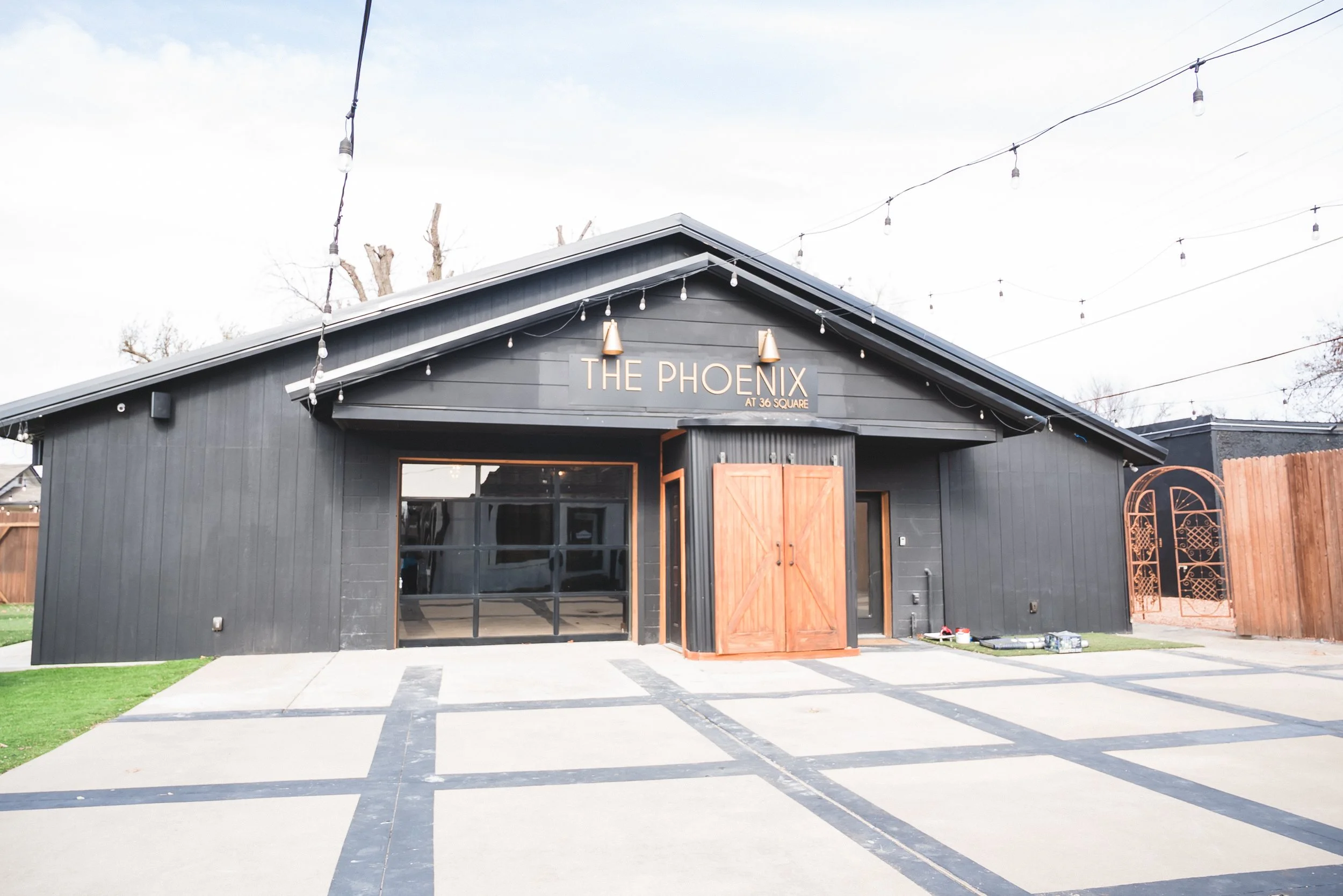 A modern black building with a sign that reads 'The Phoenix at 36 Square.' The building has a gray metal roof, string lights overhead, large windows, and a wooden double door. There is a paved area outside with some patches of grass on the sides.