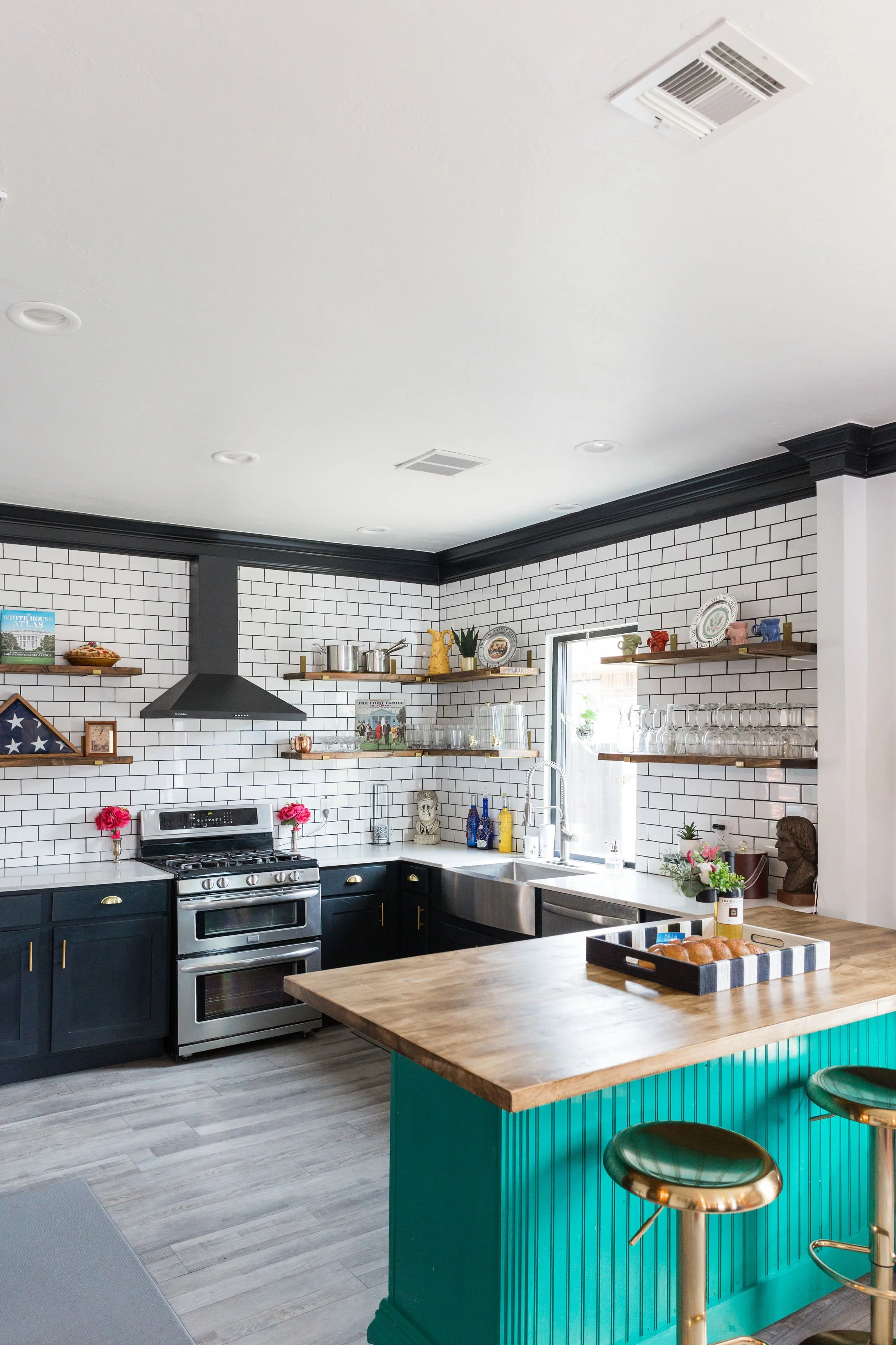 Modern kitchen with black cabinets, white subway tile backsplash, open wooden shelves, stainless steel appliances, and a green kitchen island with wooden countertop and gold stools.