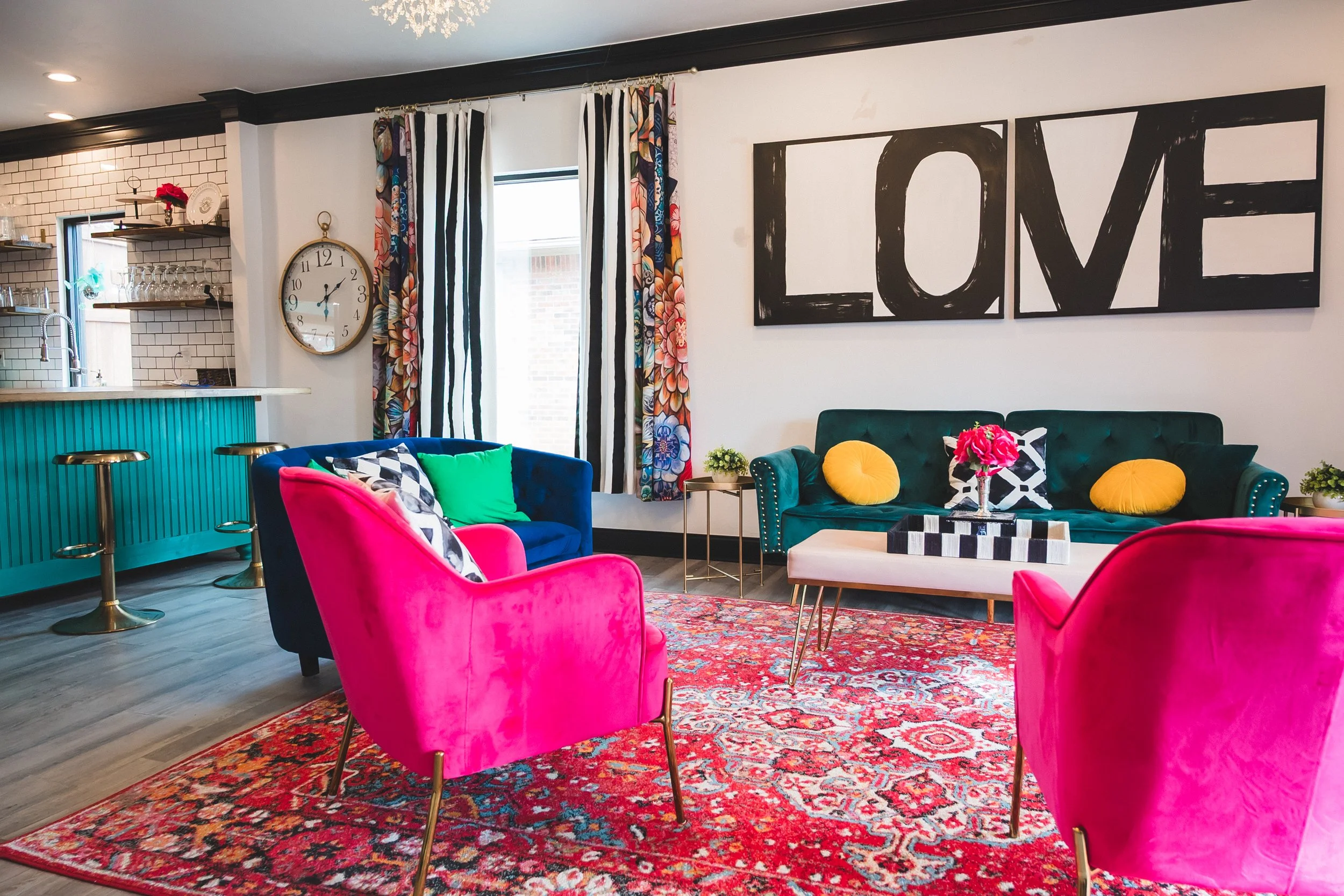 Colorful living room with a teal couch, pink armchairs, patterned curtains, a large wall art spelling LOVE, a vibrant area rug, and a small white coffee table with a flower vase.