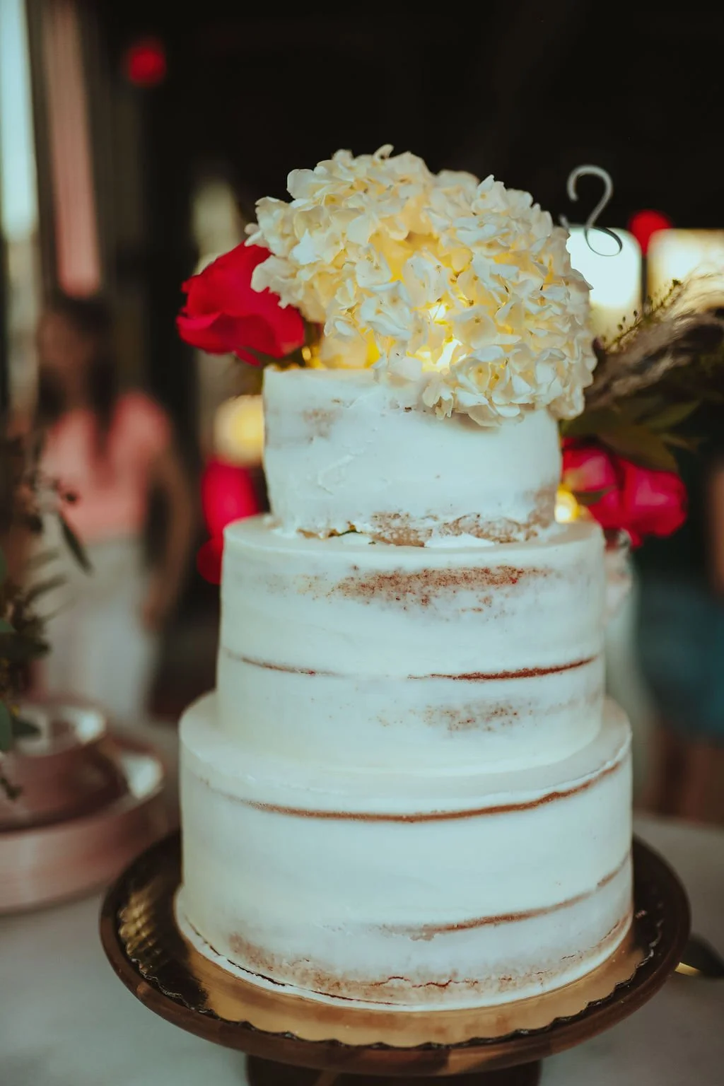 A four-tiered wedding cake with a rustic semi-naked finish, decorated with white hydrangea flowers and red roses on top, on a black cake stand.