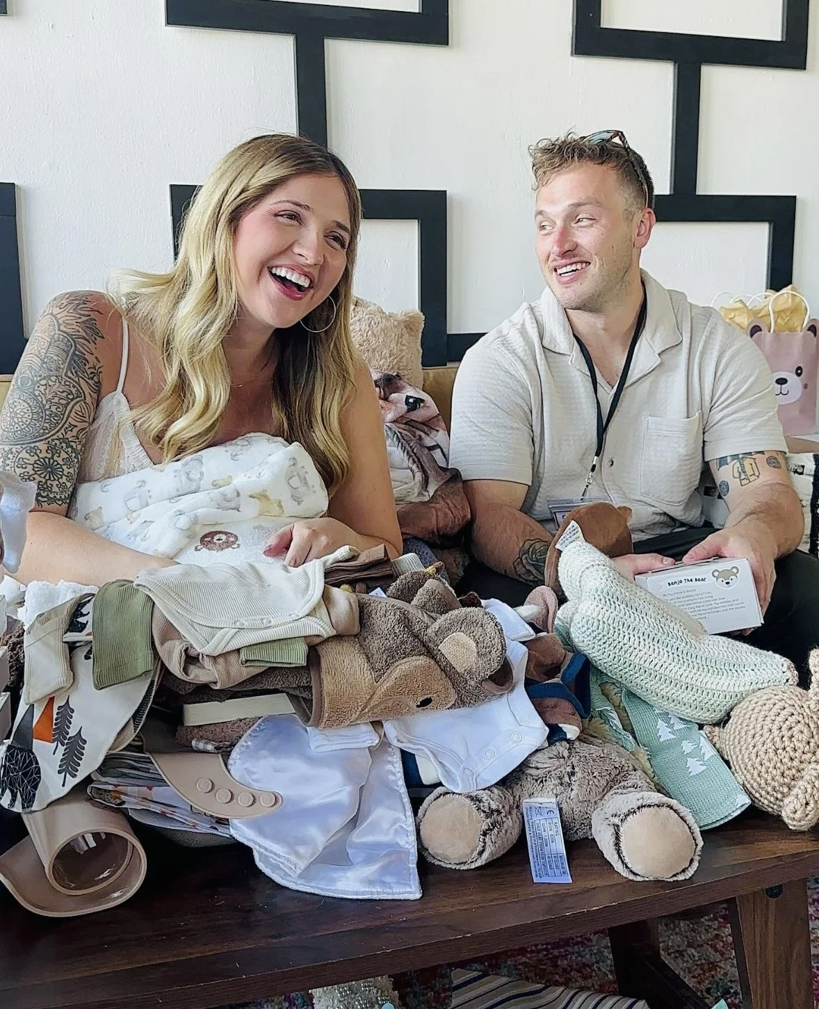 Two adults, a woman and a man, are sitting at a table filled with baby clothes and stuffed animals, smiling and engaging with each other in a bright room.