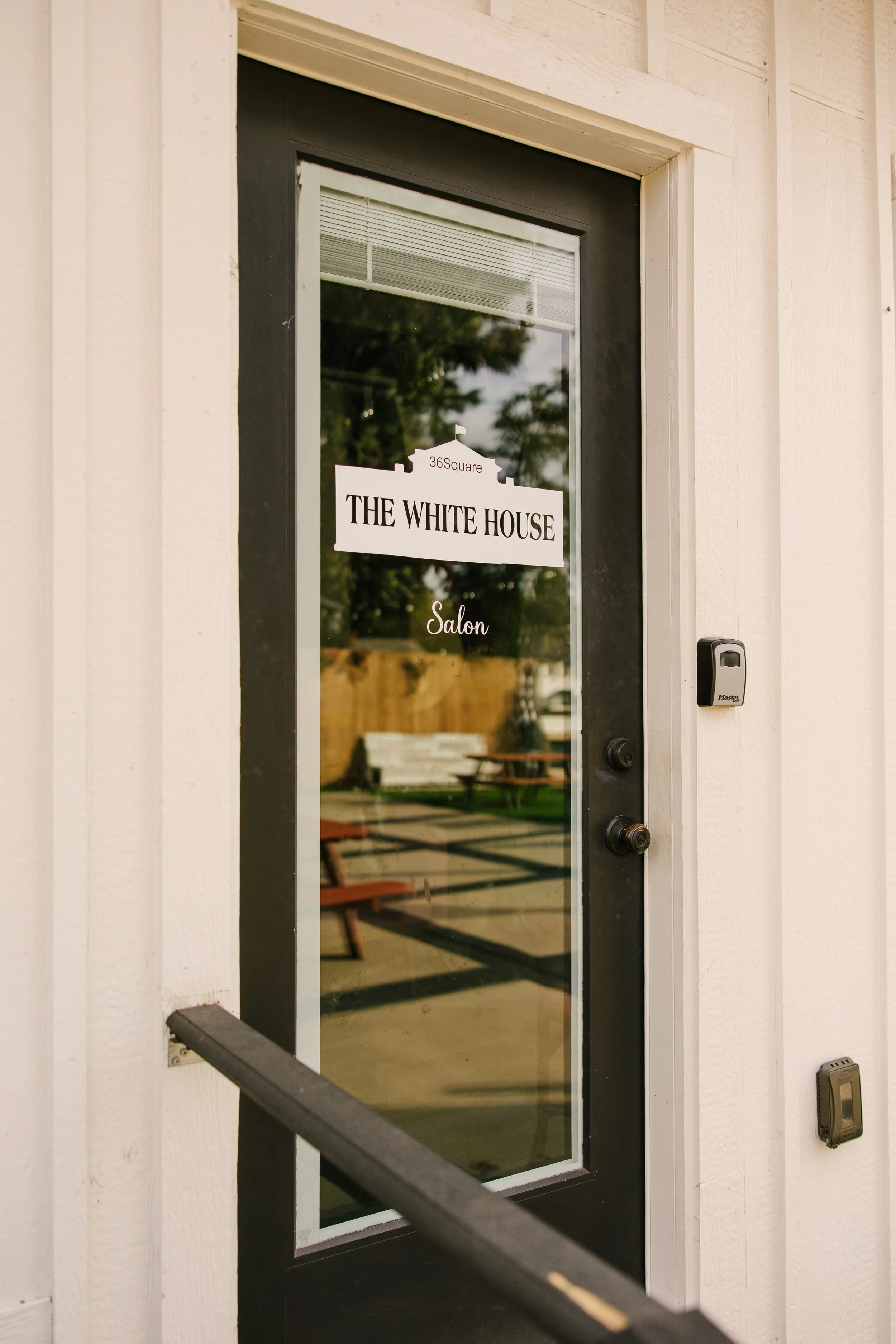 Close-up of a black glass door with signs for 'THE WHITE HOUSE' salon, reflecting outdoor seating area with picnic tables and trees.