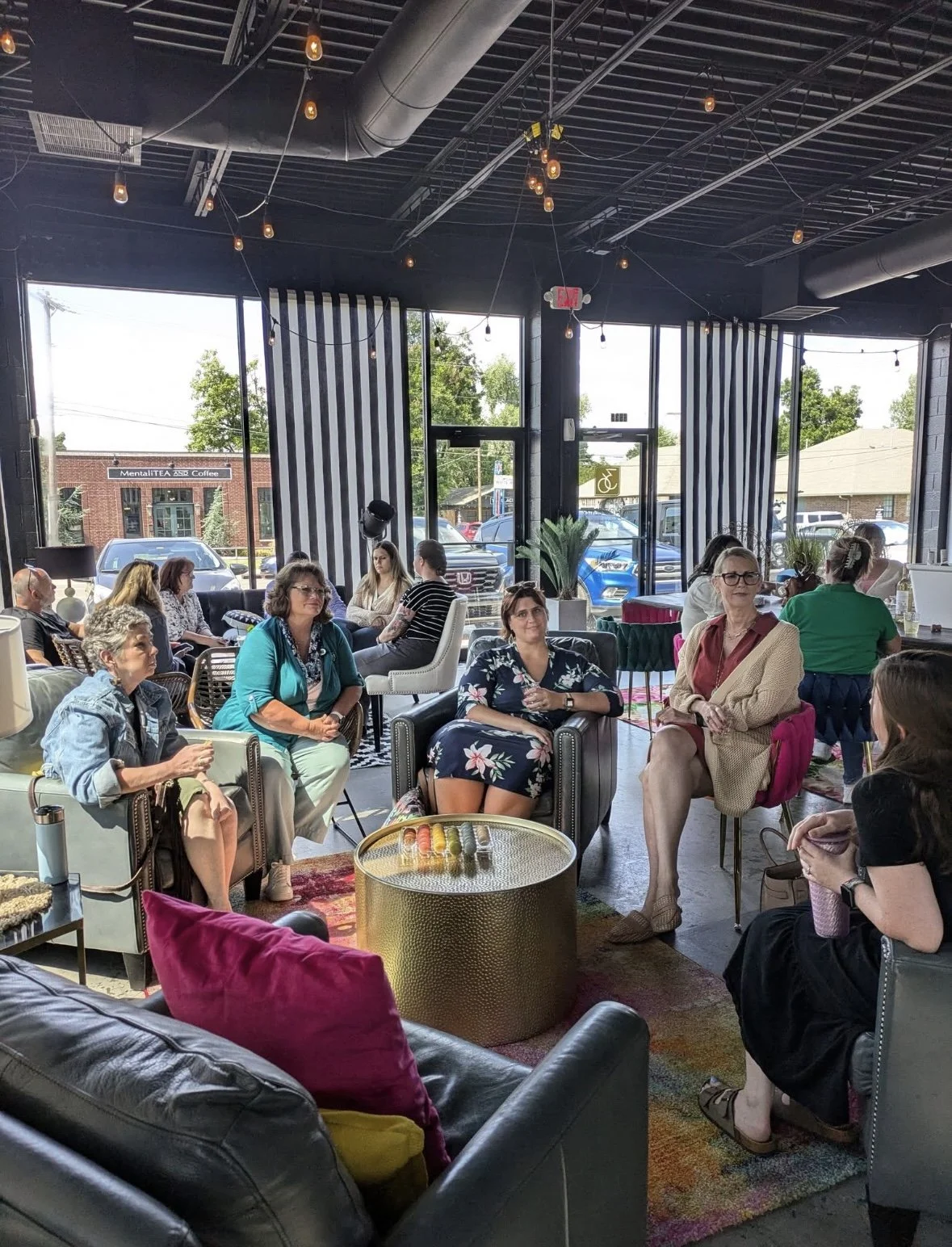 A group of women sitting in a circle inside a modern coffee shop during the day, with large windows showing parked cars and a street outside, decorated with string lights and potted plants.