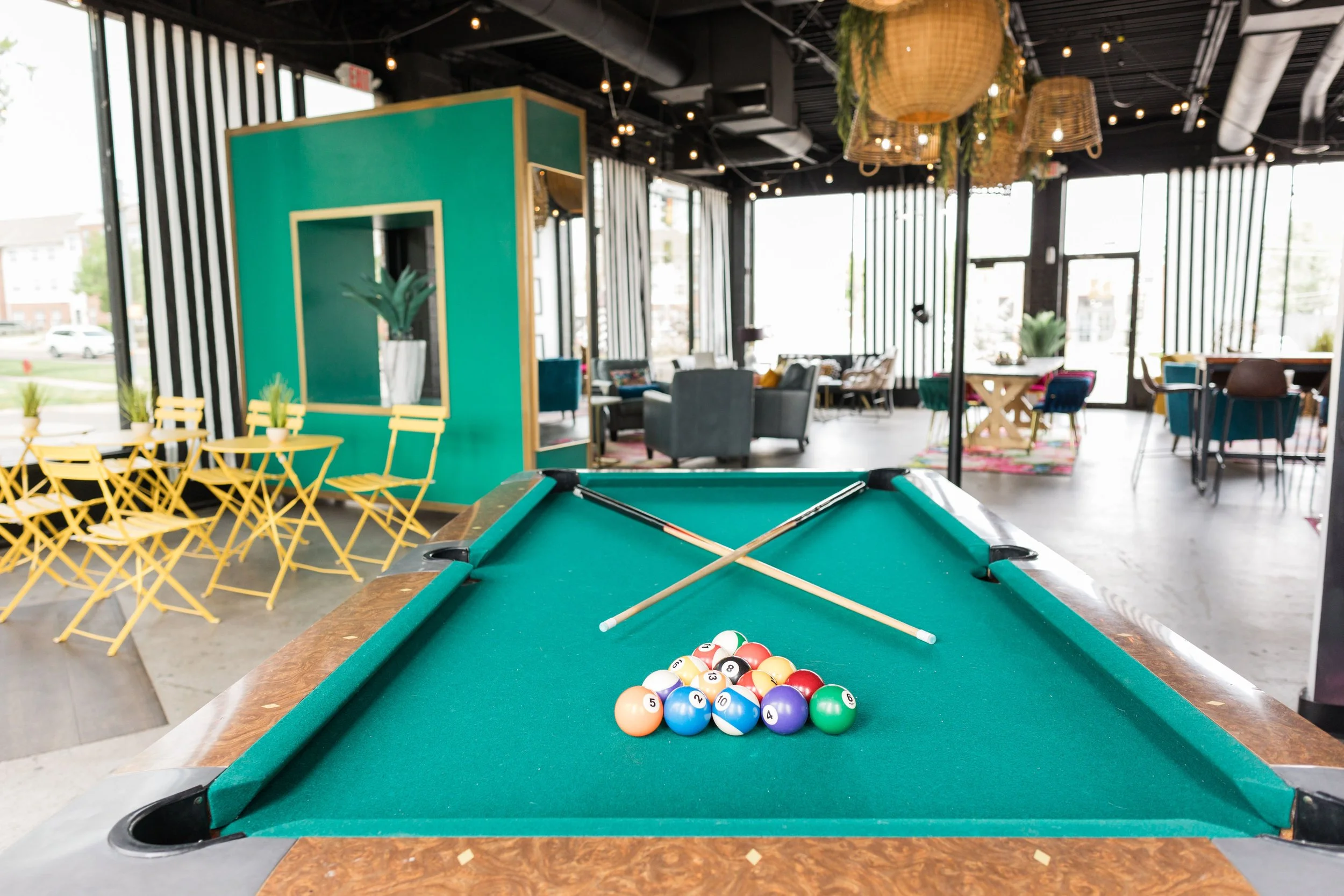 Pool table with balls racked and two cues crossed on it inside a modern lounge or cafe with yellow chairs and large windows.