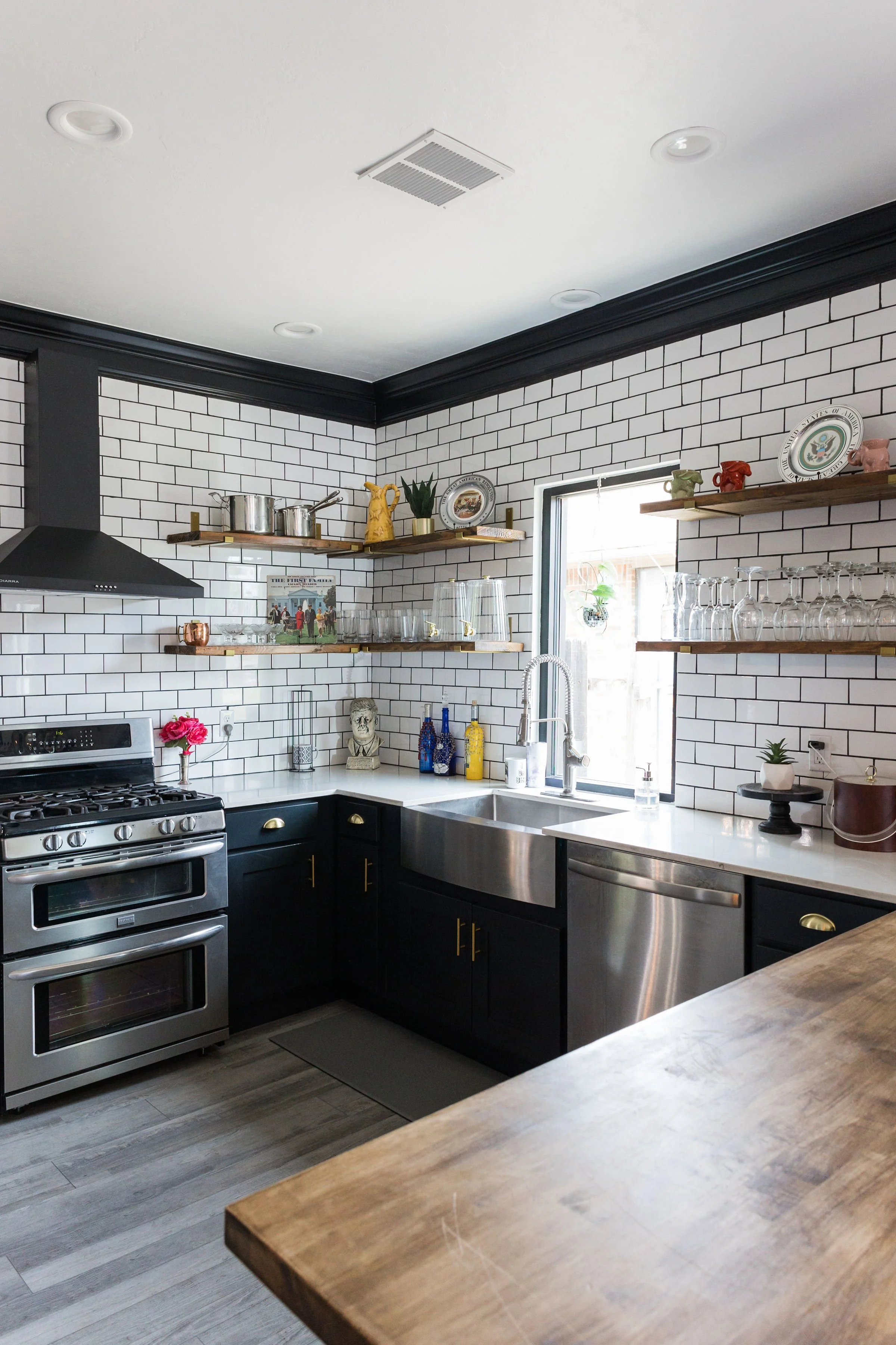 Modern kitchen with white subway tile walls, black cabinets with brass handles, stainless steel appliances, wooden open shelves with various decorative items and glassware, and a window above the sink.