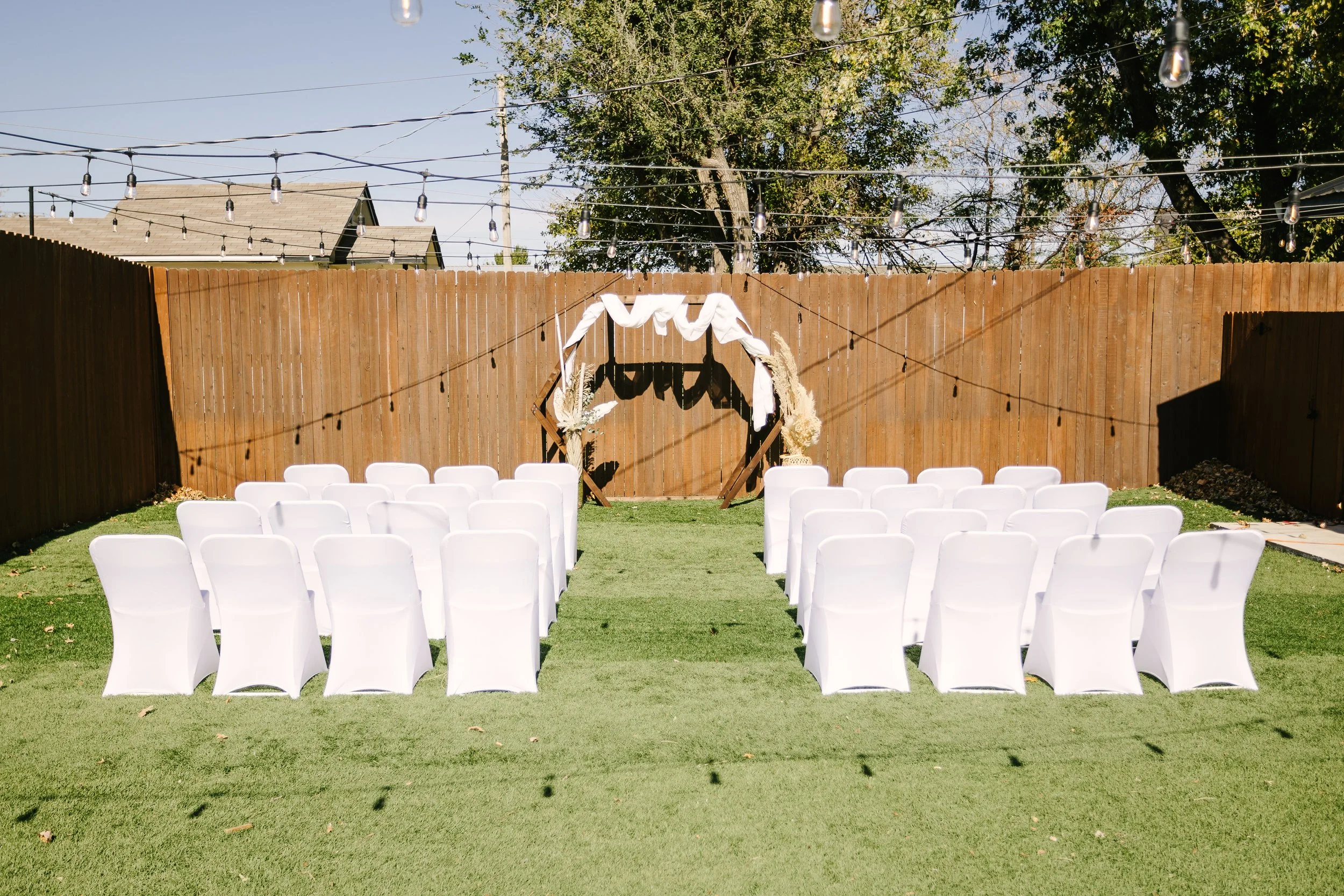 Outdoor wedding setup with white chairs, a decorative arch with fabric, and string lights on a grassy yard enclosed by a wooden fence.