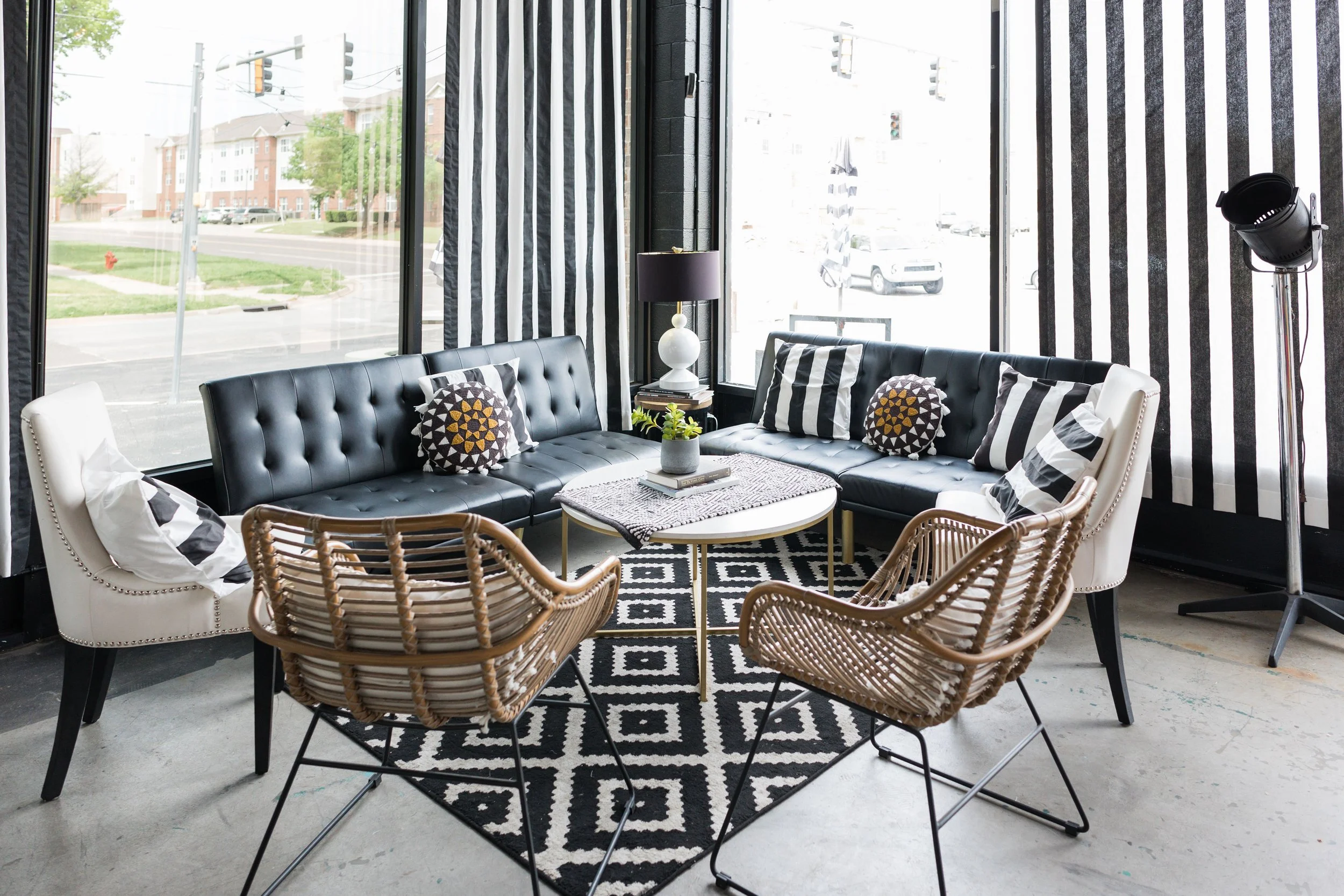 Modern living room with black and white striped curtains, black leather couch, white armchairs, a round coffee table with a plant and books, and large windows showing a street view outside.