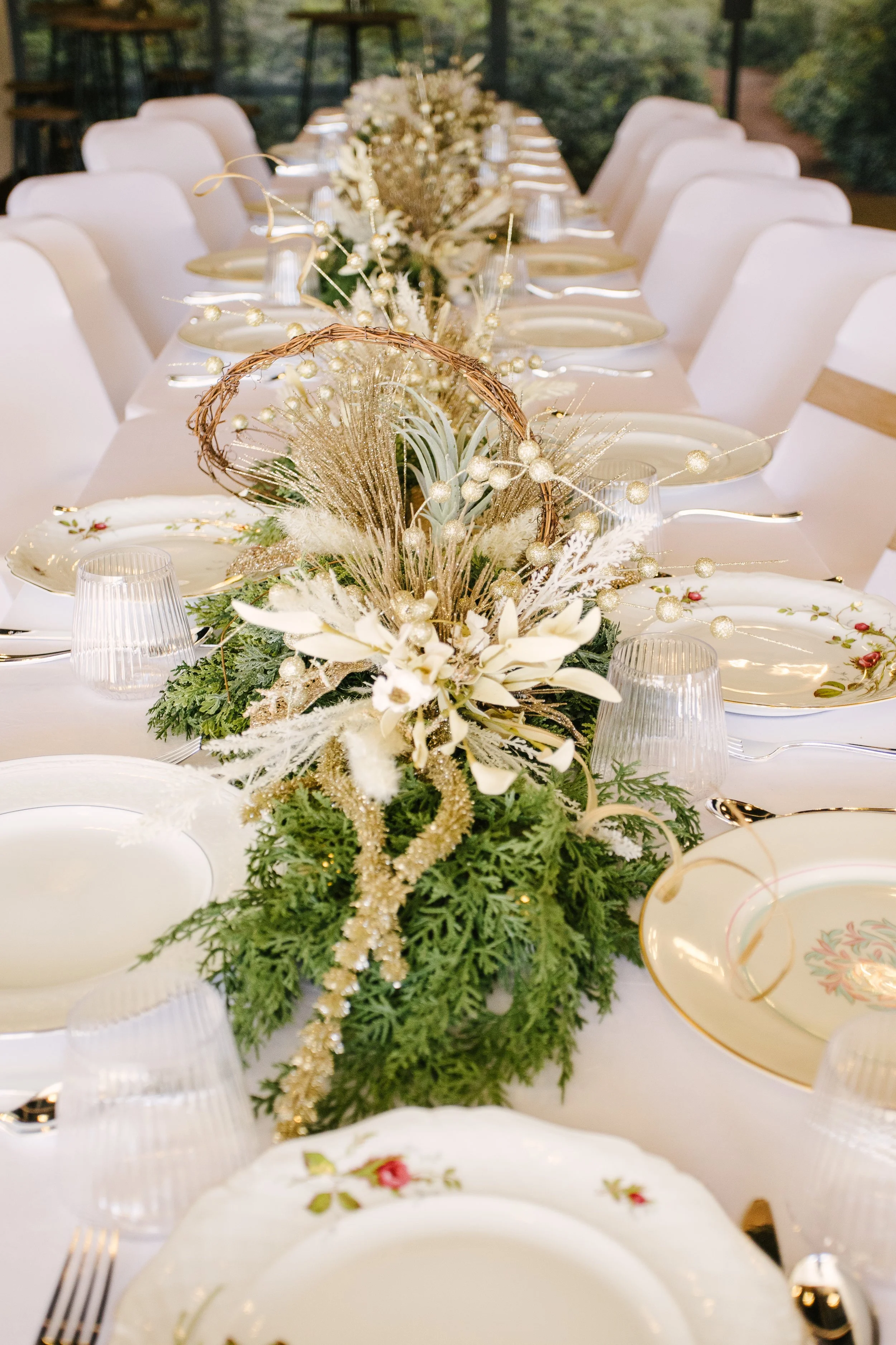 Elegant dining table decorated with a white tablecloth, floral china, and a gold-themed centerpiece featuring white flowers, greenery, and gold accents for a festive occasion.