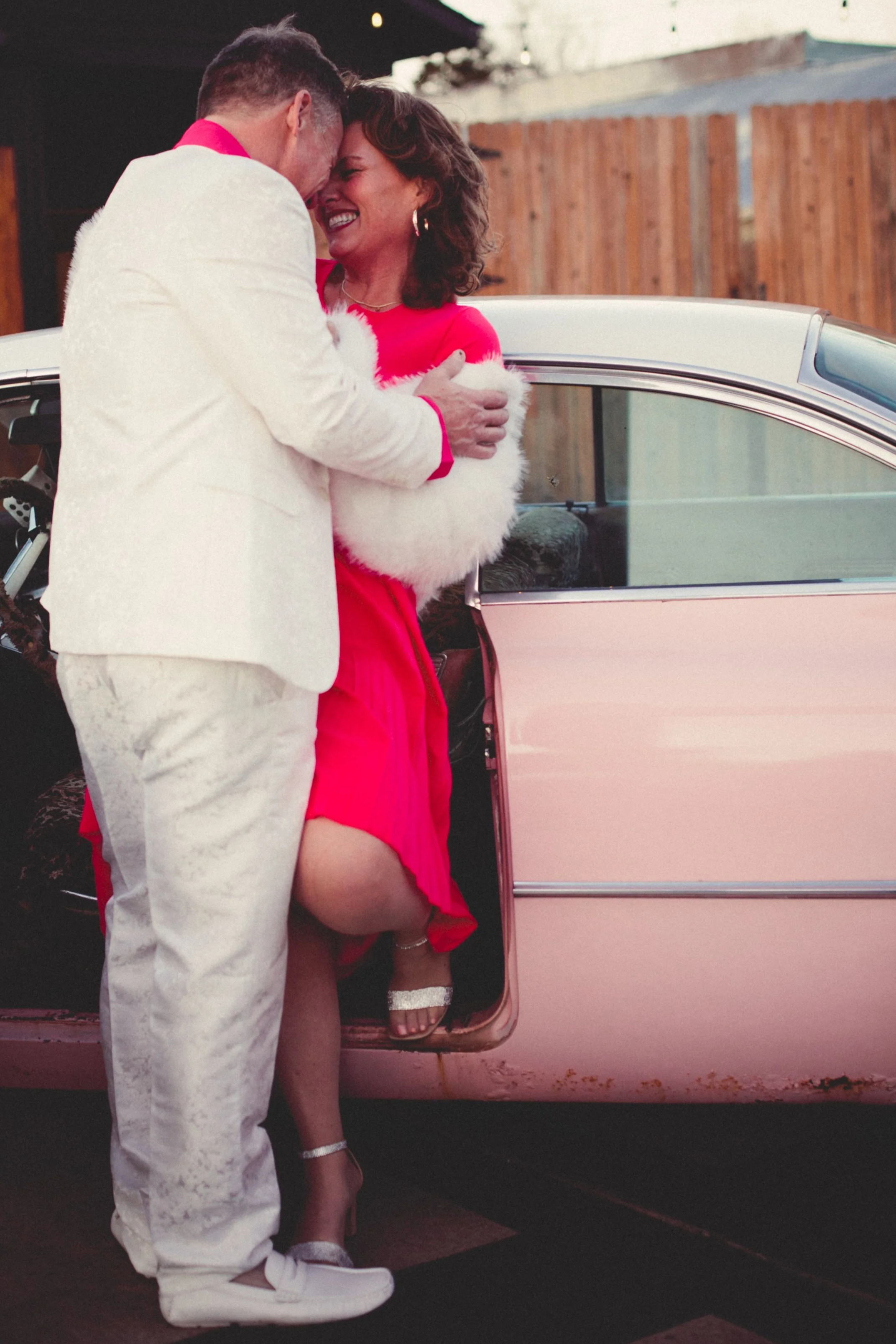 A couple dressed in festive holiday attire sharing a joyful moment outside a vintage pink car.