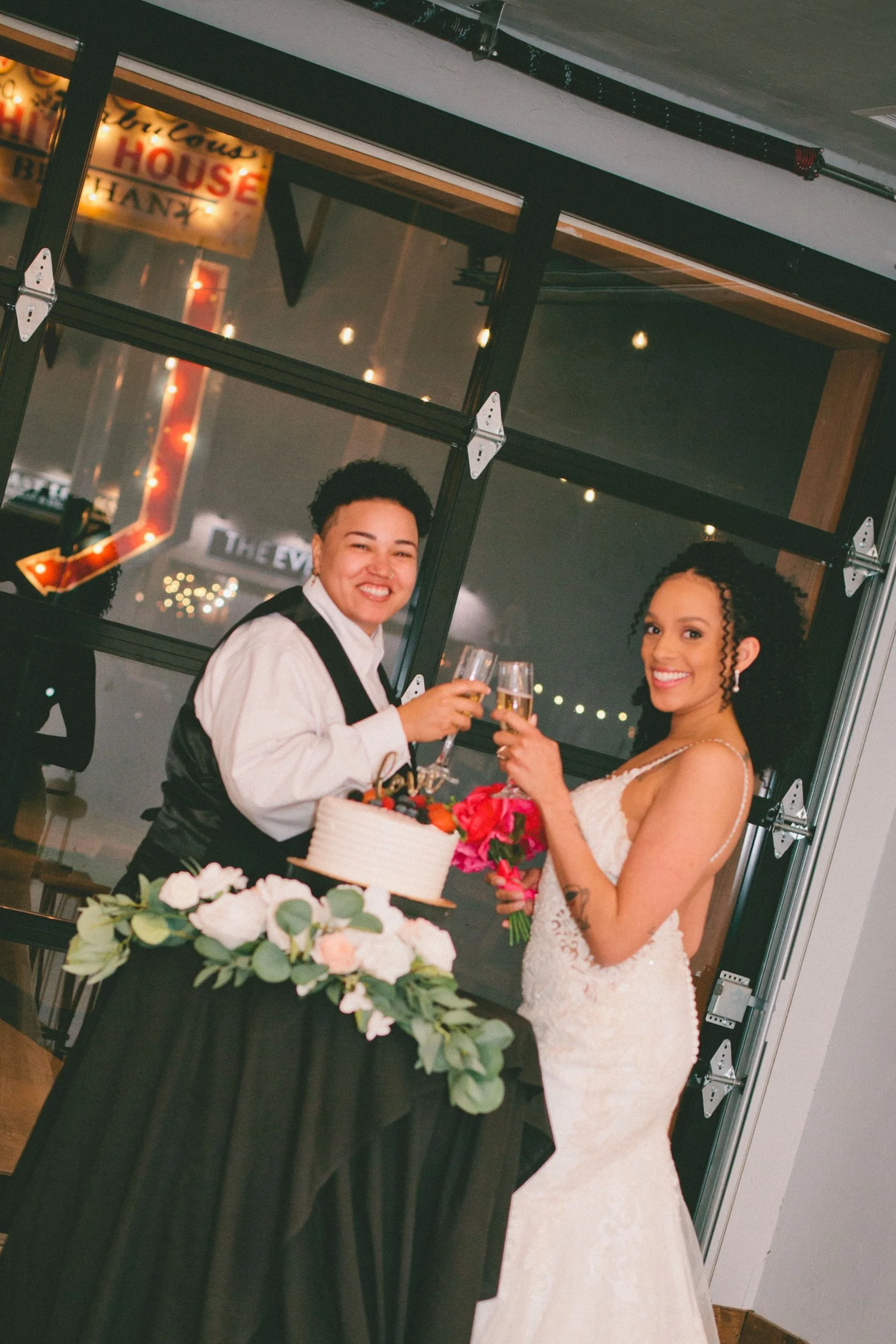 Two women, one in a wedding dress and the other in a tuxedo, are celebrating with champagne at a wedding reception, standing next to a table with a cake and floral decorations.