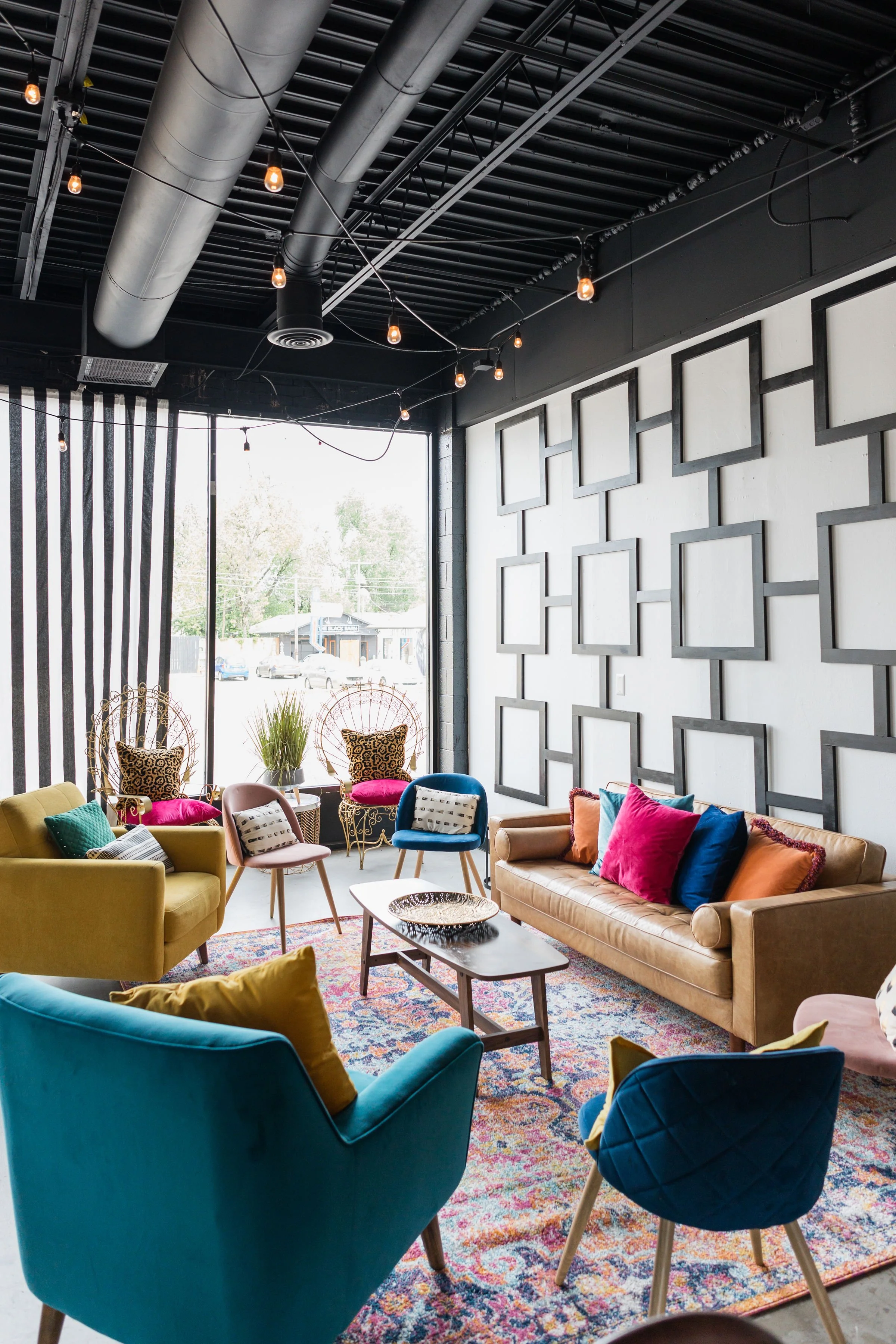 Colorful modern lounge with eclectic furniture including velvet armchairs, a leather sofa, patterned pillows, a wooden coffee table, and a vibrant rug. Large window with vertical striped curtains, decorative wall with geometric frames, and string lig