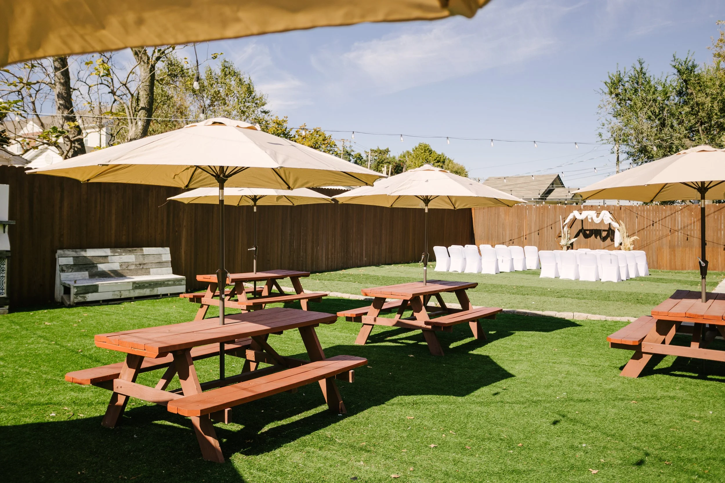 Outdoor setup with picnic tables and umbrellas, a row of white chairs, and a decorative arch for a wedding or event in a backyard.