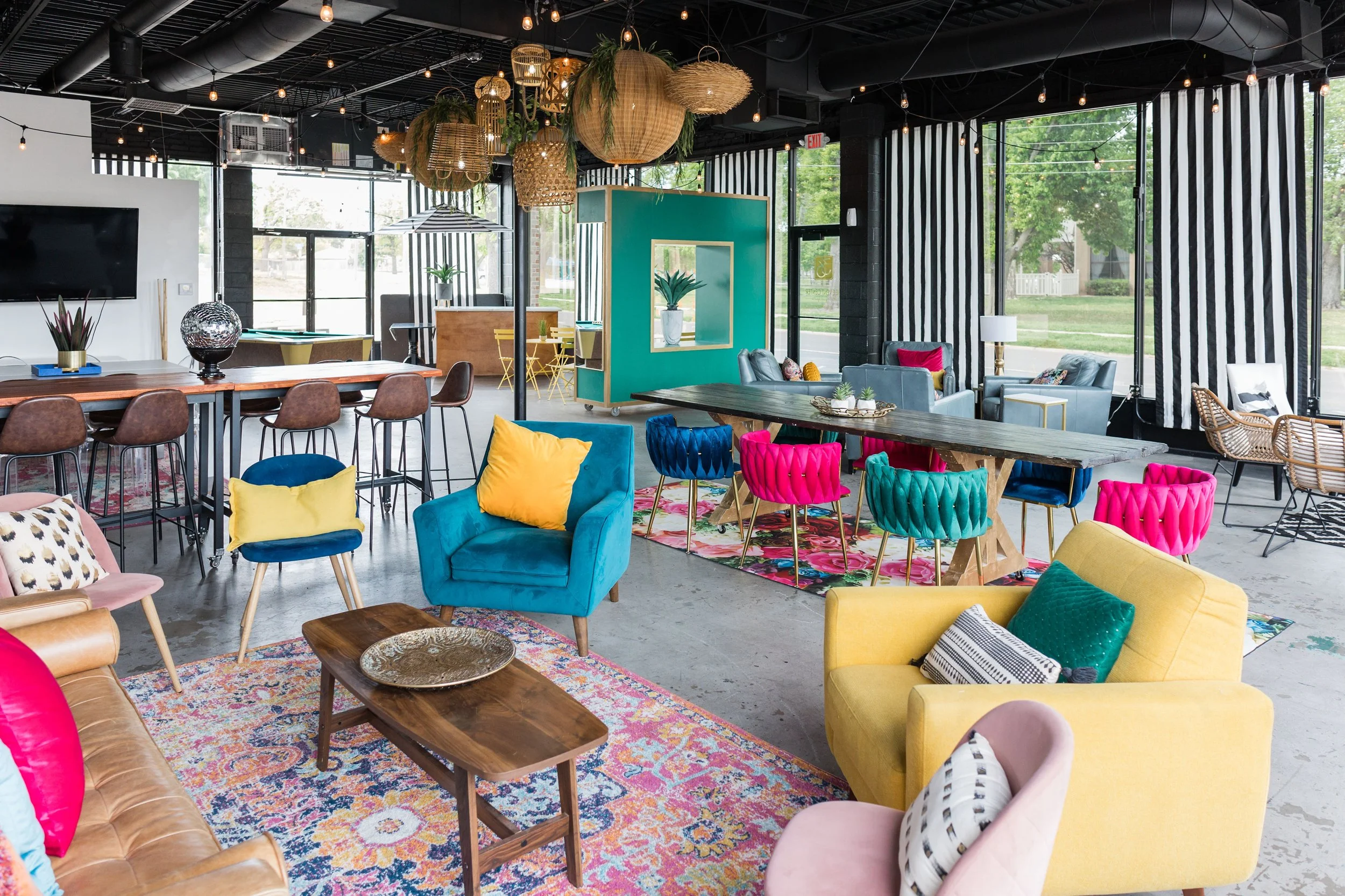 Colorful, modern lounge with various sofas, armchairs, and tables, decorated with vibrant cushions and rugs, large windows, and warm lighting.