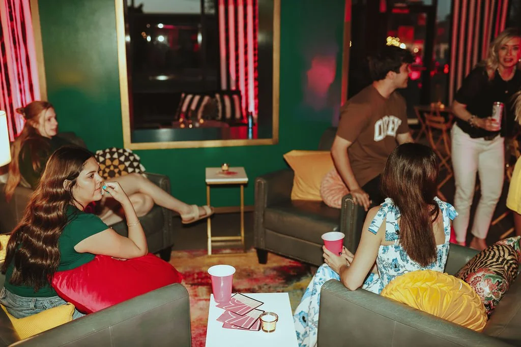 Young women and a young man socializing in a dimly lit lounge or bar with colorful walls, some sitting on couches holding drinks, others standing and chatting.