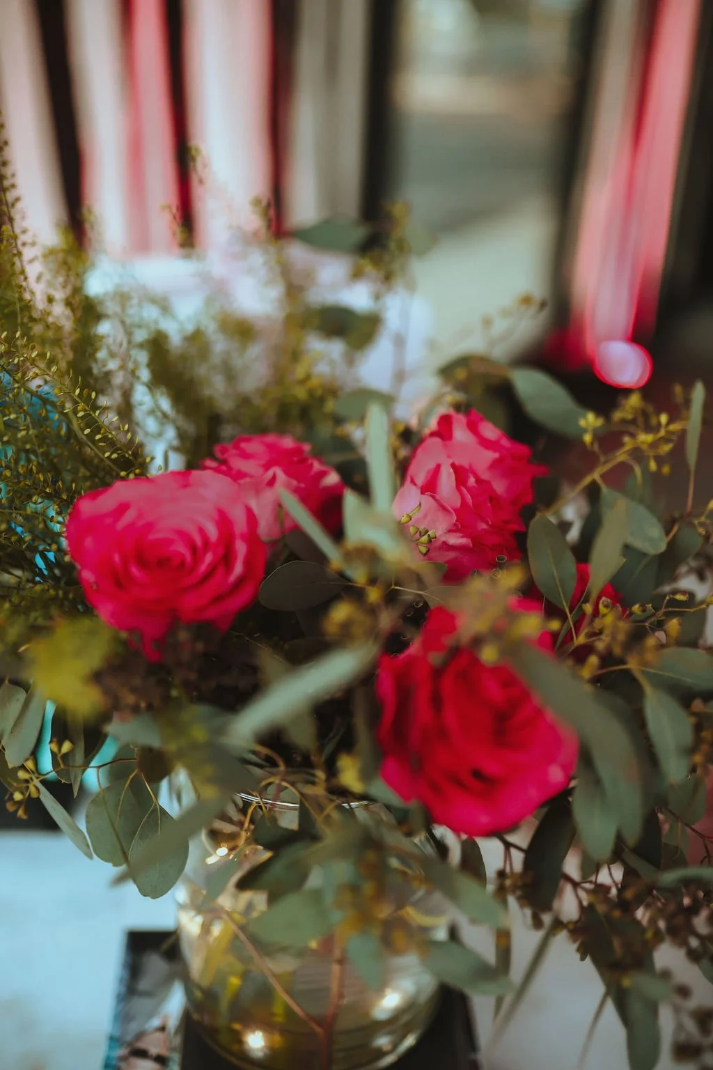 A close-up of a bouquet of red roses and green foliage in a glass vase, with a blurry background featuring curtains and a wall with a circular light.