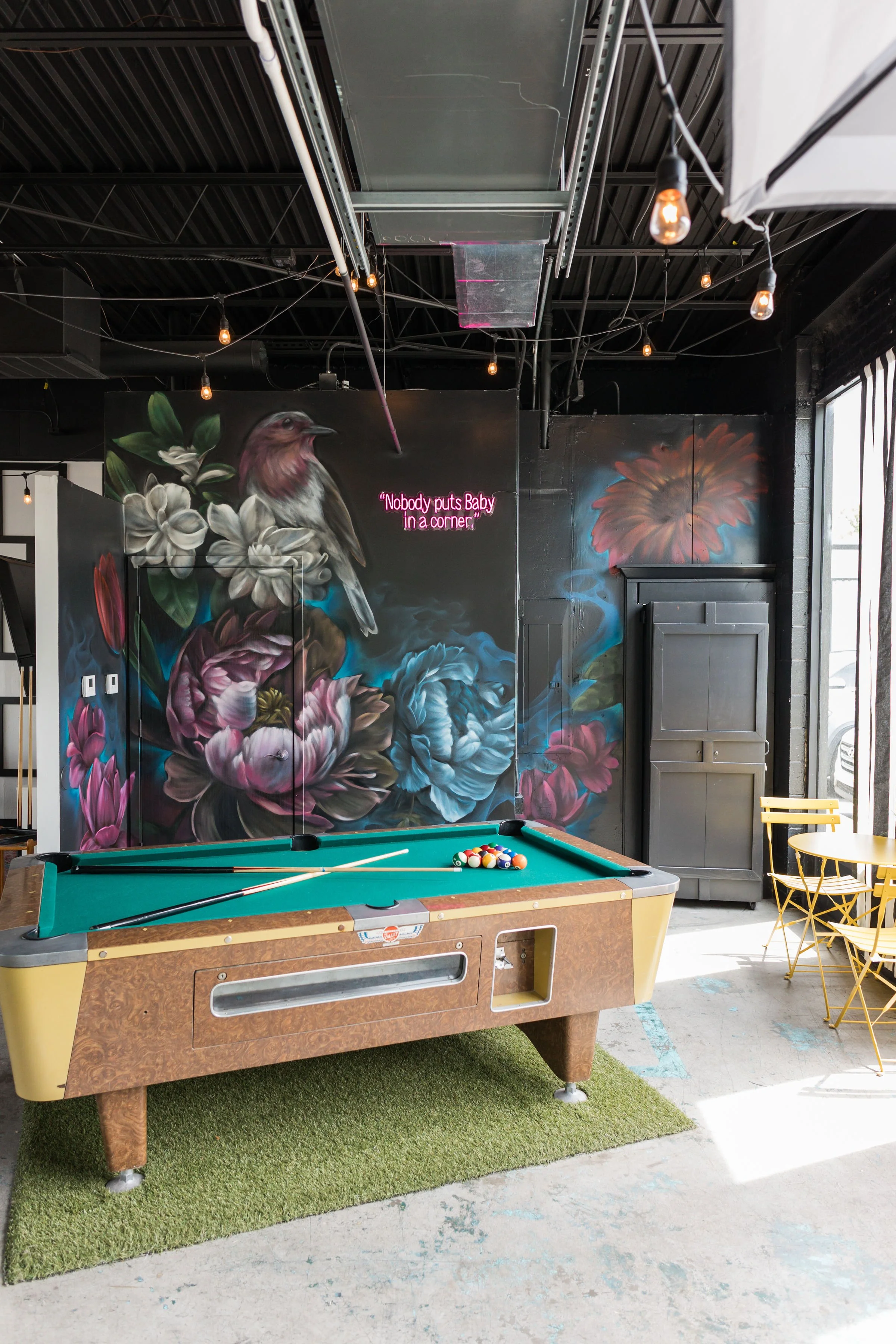 An indoor scene with a vintage pool table, a mural of large flowers and a bird on the wall, and a neon sign that reads 'Nobody puts Baby in a corner.' There is a yellow table and chairs to the right and sunlight coming through a window.