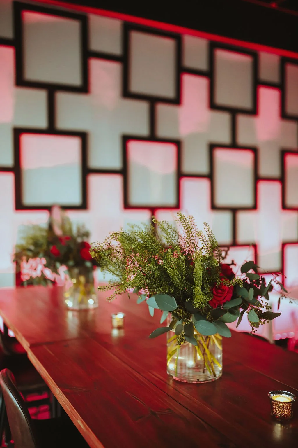Vase with pink roses and green foliage on a wooden table, with decorative candles and a geometric patterned wall in the background.
