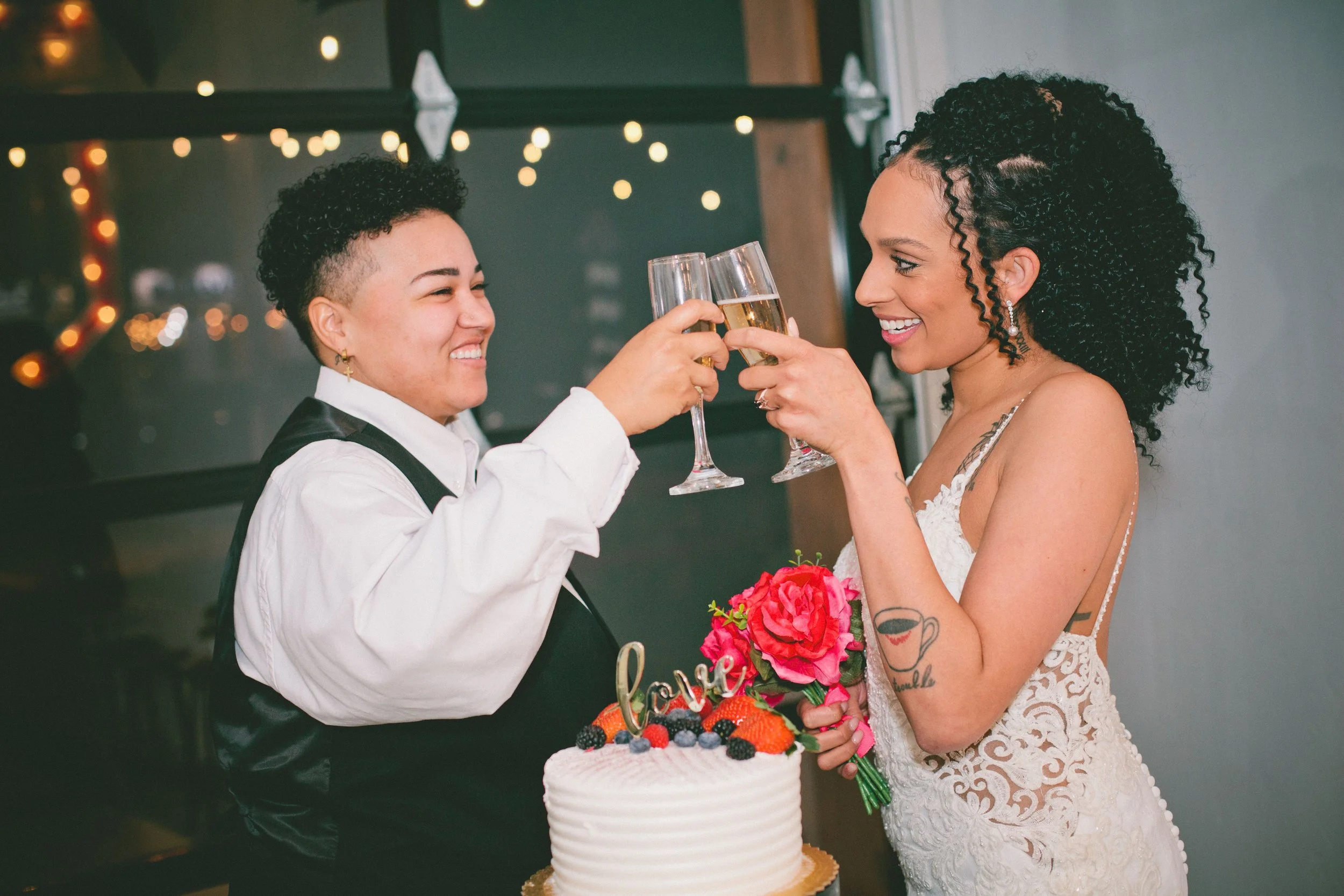 Two women celebrating with champagne toast at a wedding or party, one holding a bouquet and the other holding a cake with fruit and a "love" topper.