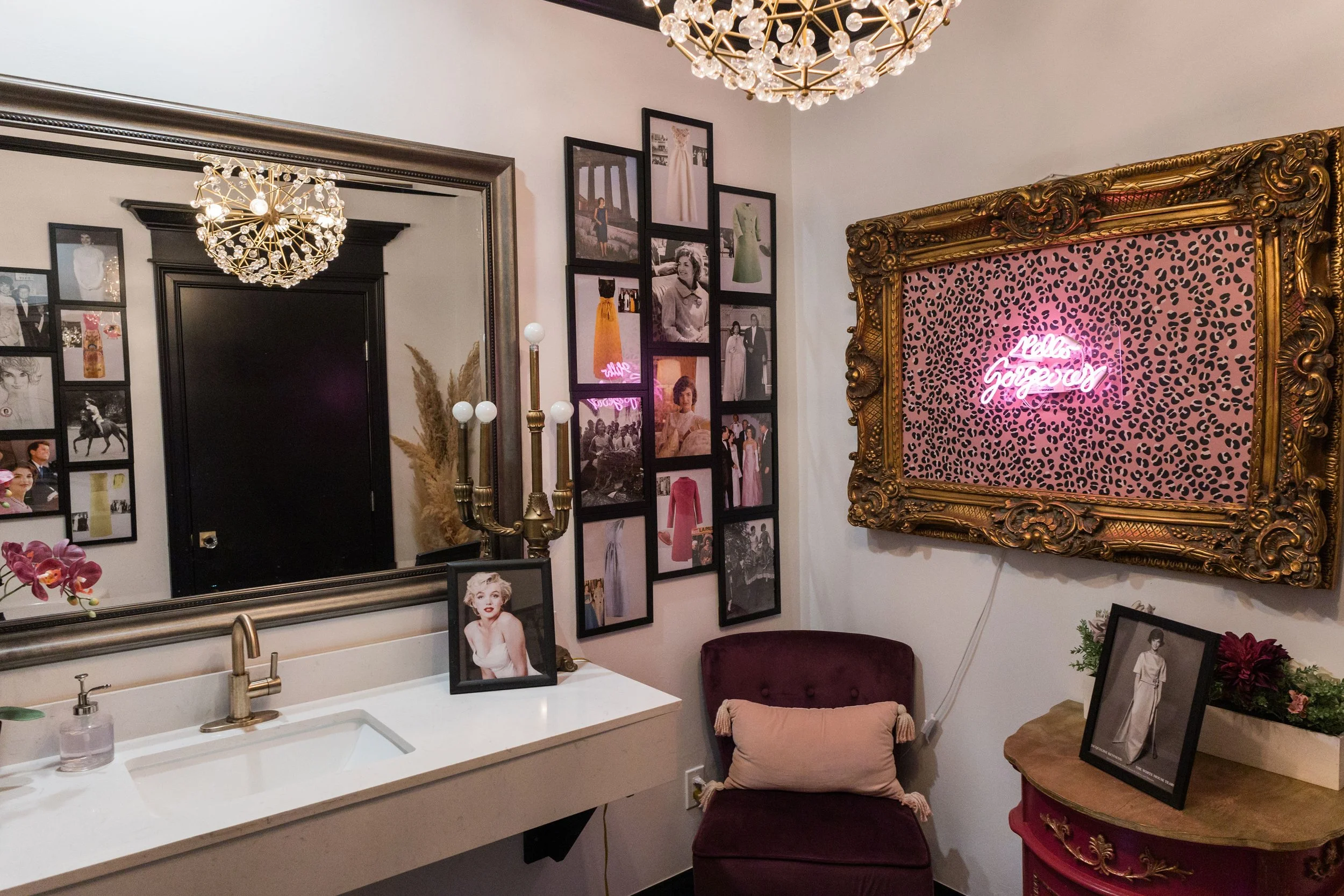 Decorative room with a large mirror, framed photos, vintage lighting, a burgundy chair with a beige pillow, a pink leopard print neon sign saying 'Girls Gorgeous', and a small ornate table with flowers.