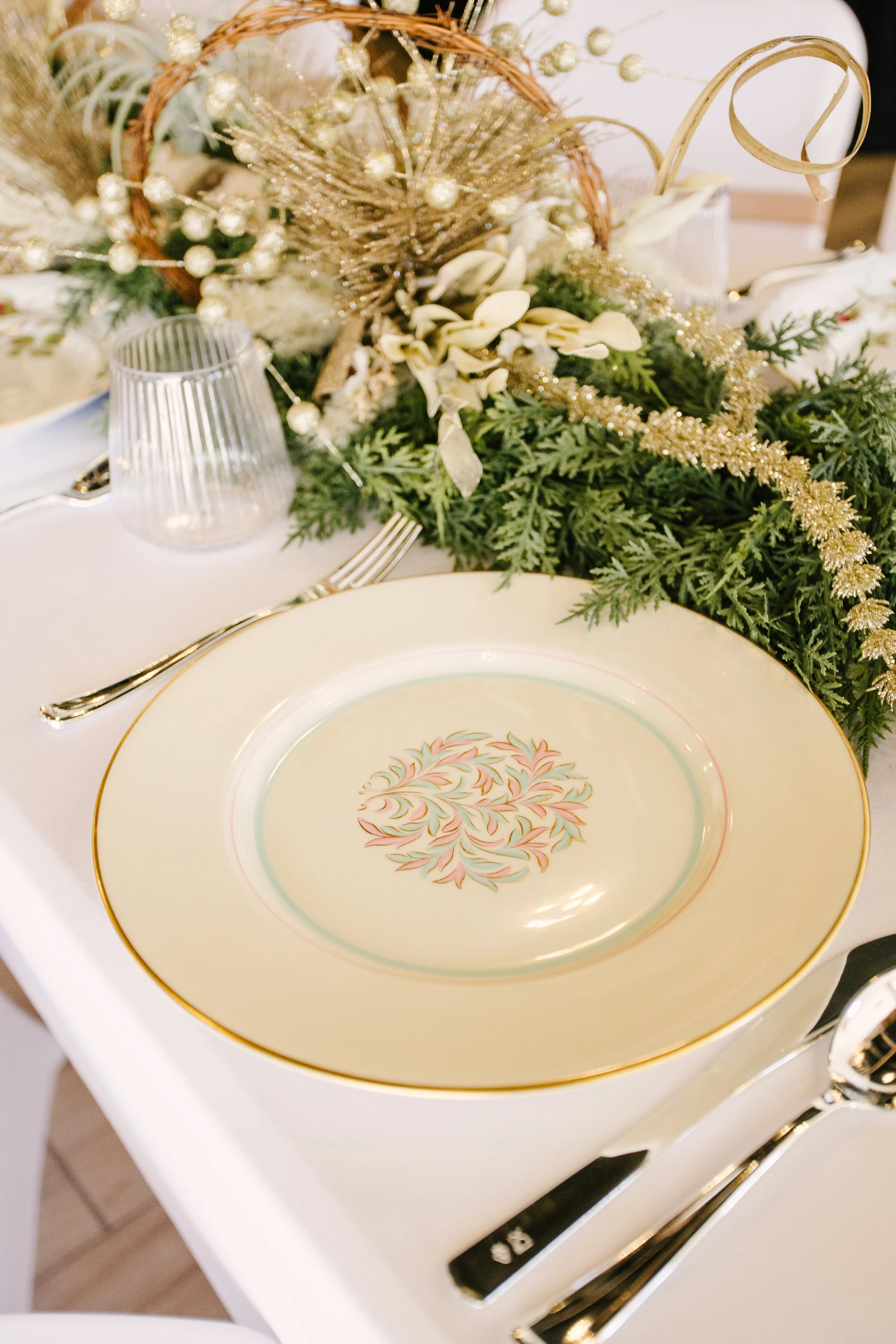 A decorated holiday table with a white plate featuring a gold trim and colorful floral pattern, surrounded by silverware, glasses, and a festive green and gold centerpiece.