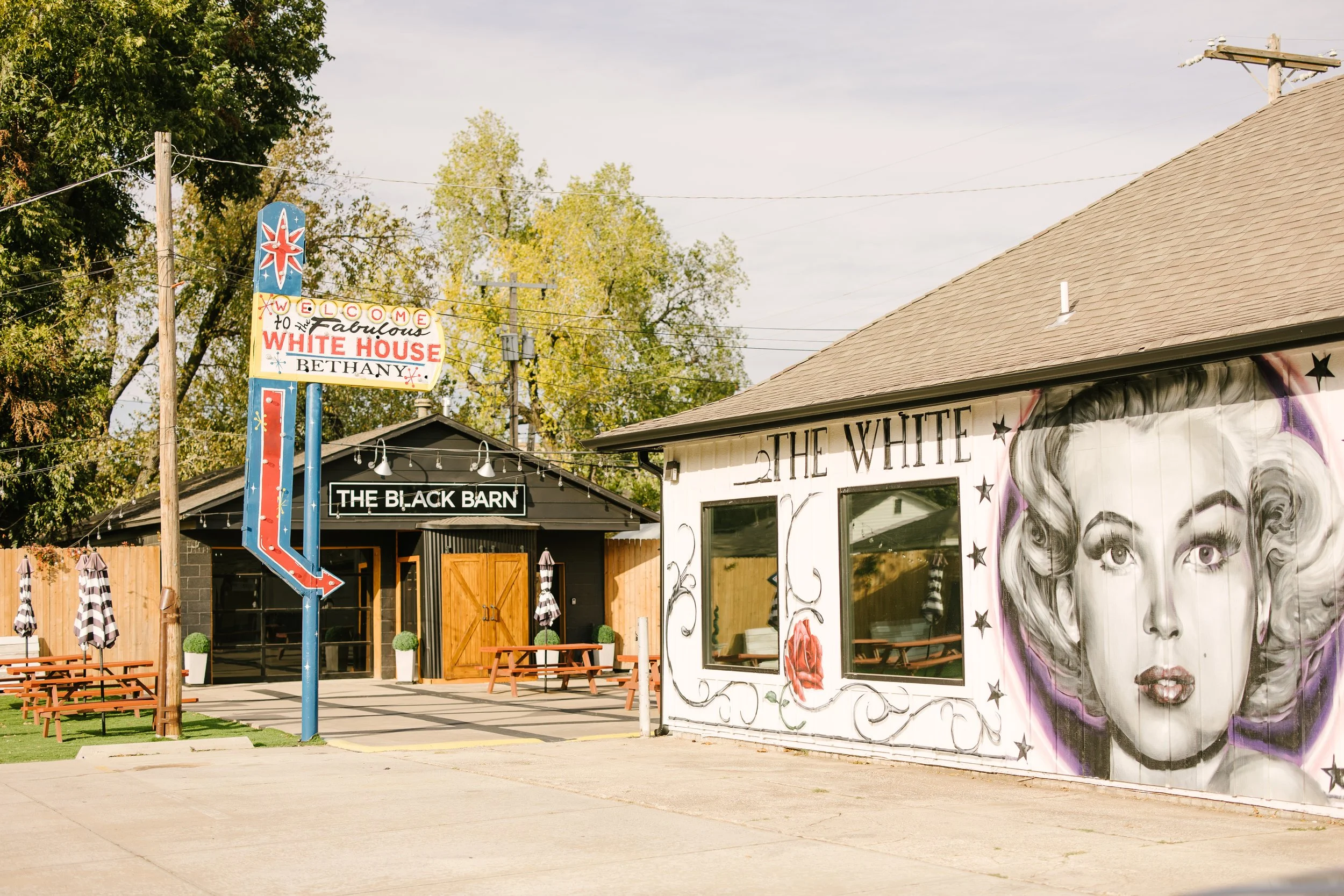 Street view of two buildings: one has a mural of a woman's face with styled hair, and the other has a sign that reads 'Welcome to the fabulous White House.' There is also a smaller building named 'The Black Barn' with picnic tables, umbrellas, and po