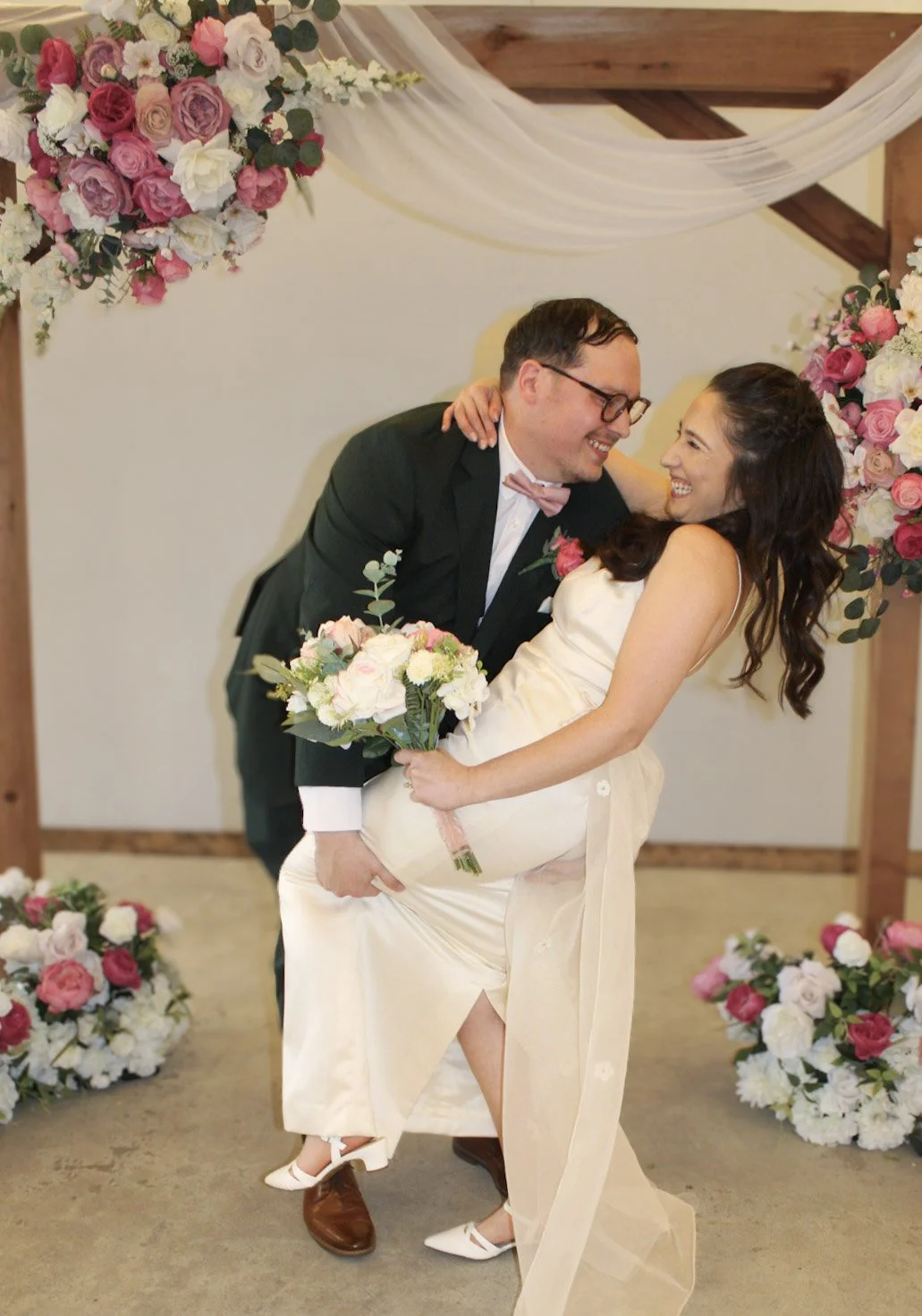 A happy bride and groom share a dance at their wedding, surrounded by pink and white floral arrangements and draped fabric.