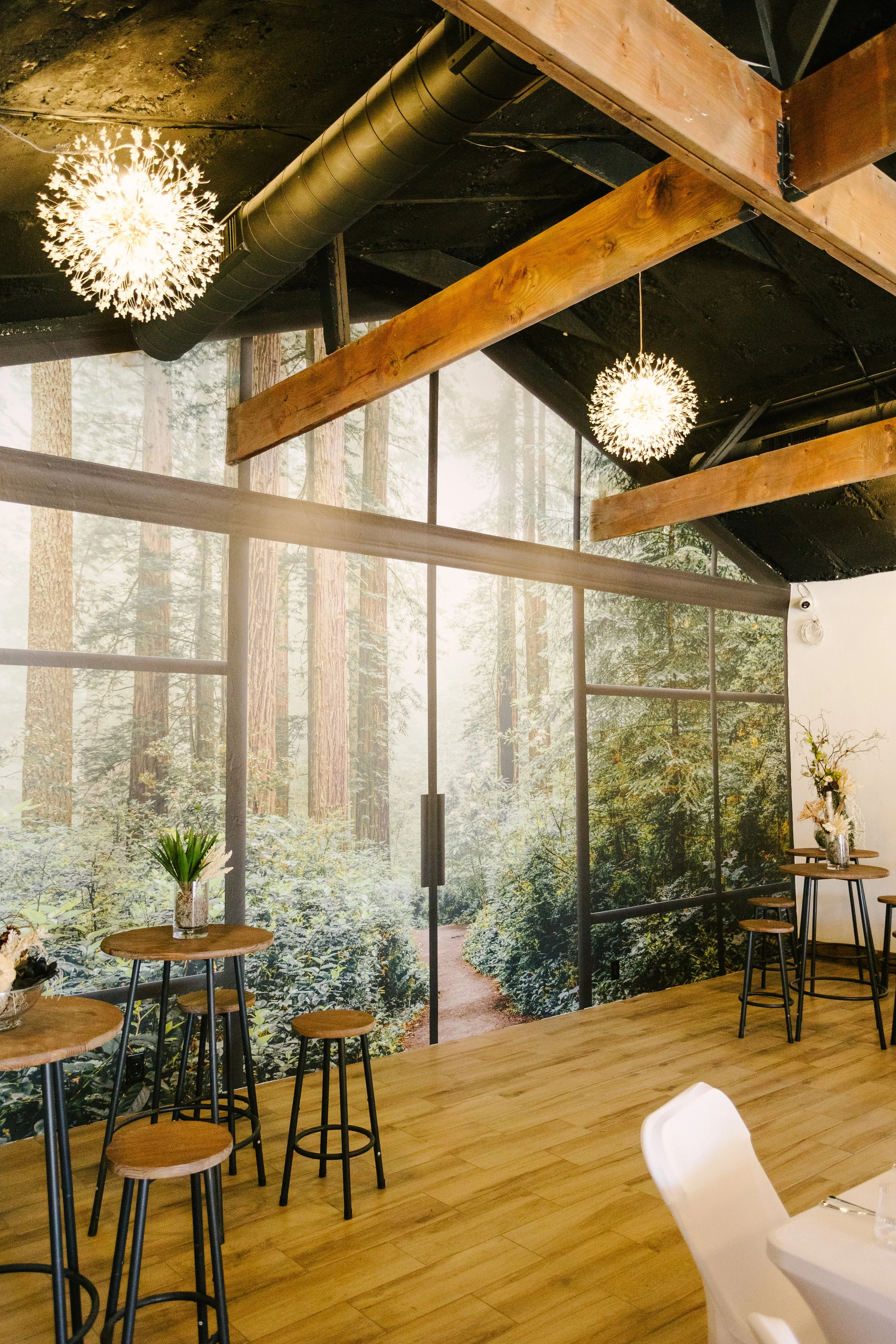 Interior of a restaurant with a large window mural of a forest, wooden tables and stools, and hanging light fixtures.