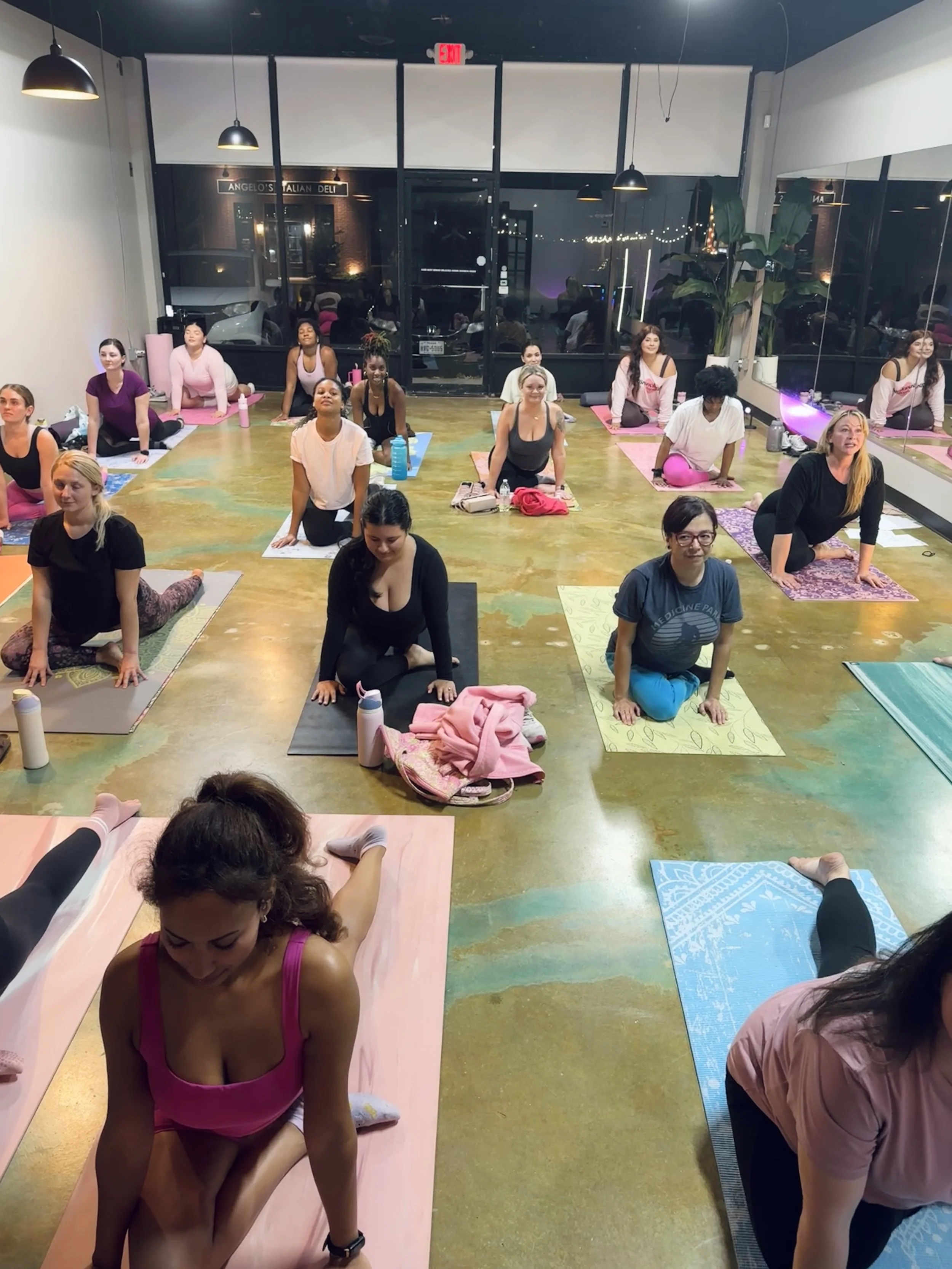 A group of women participating in a yoga class in a fitness studio. They are performing a yoga pose on colorful mats, some with water bottles and personal items nearby. The studio has black pendant lights, large windows with a view outside, and a mir