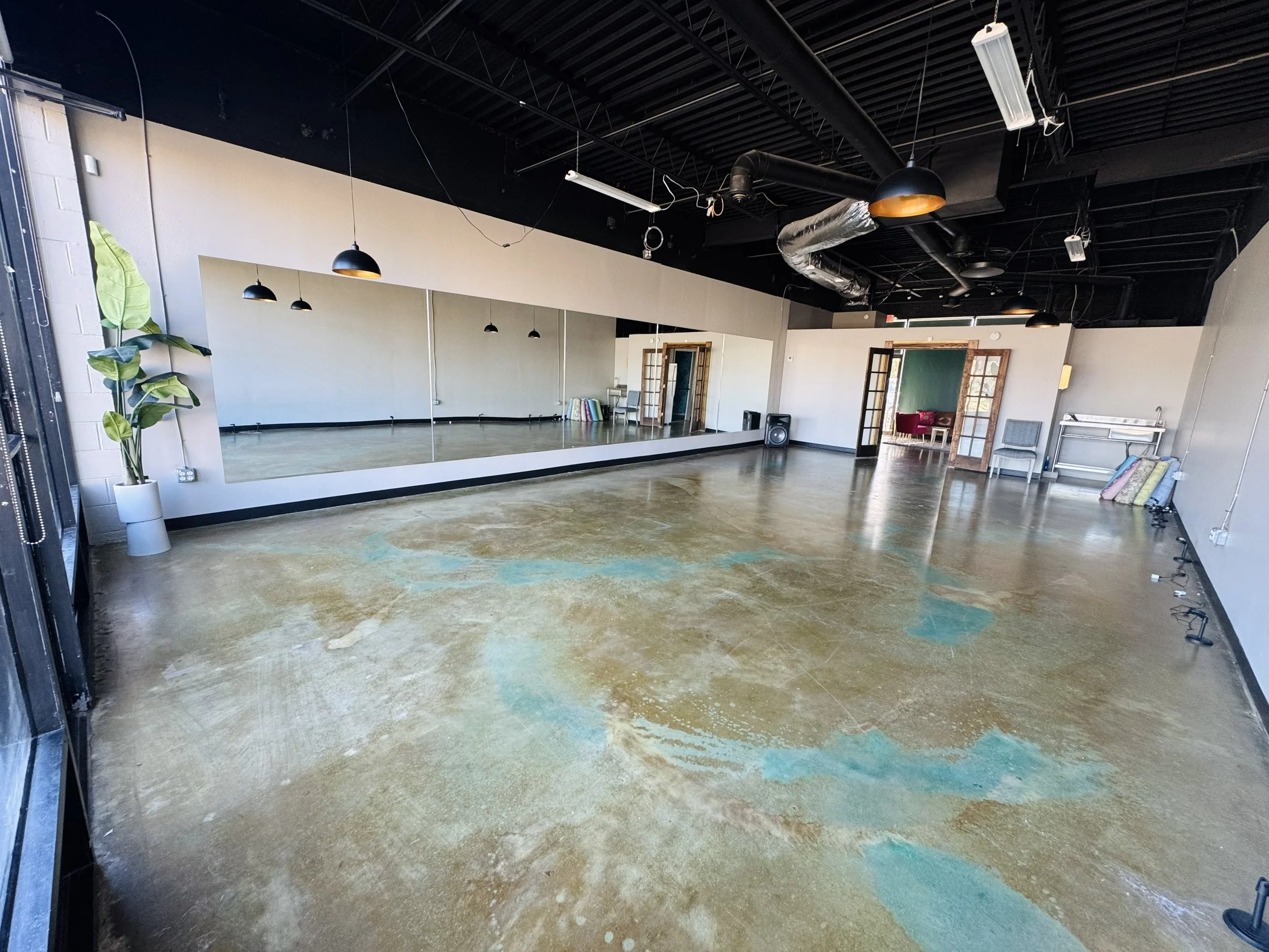Empty dance or yoga studio with large mirror, ballet barre, and hardwood floors with blue paint stains, exposed ceiling with lighting, doors leading to another room, and minimal furniture.