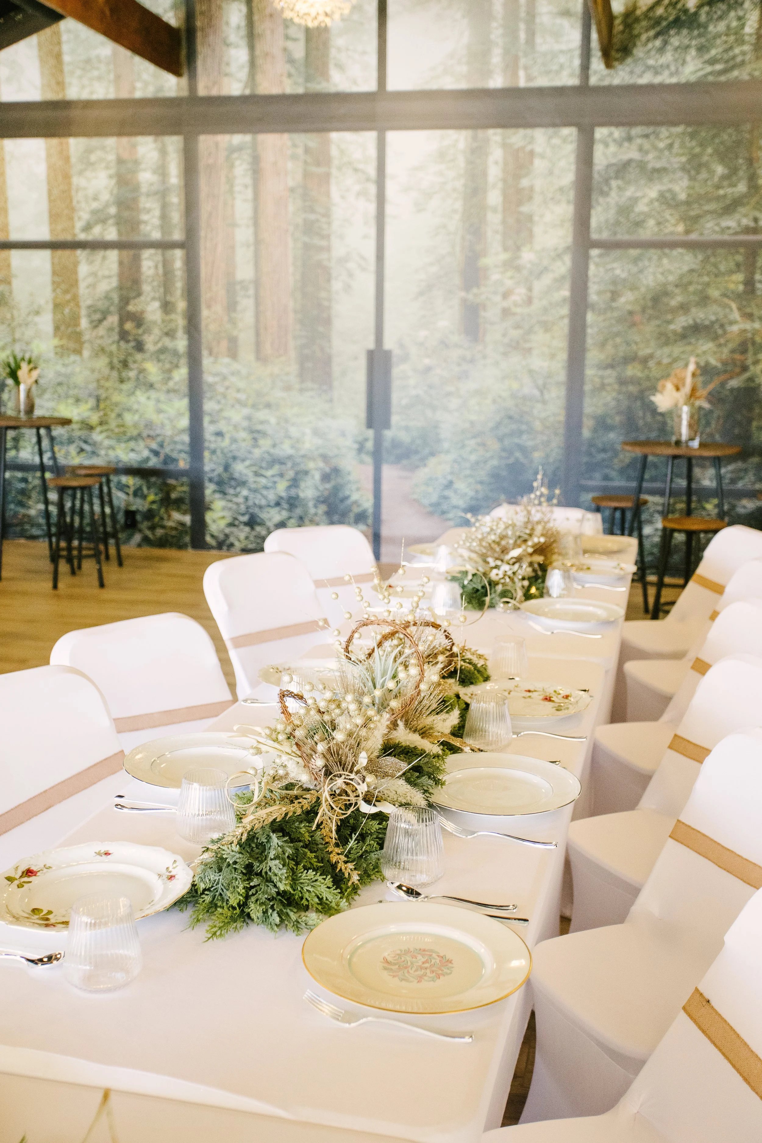 A decorated dining table set for a celebration with white chairs, festive tableware, and floral centerpieces in a room with a forest mural on the wall.