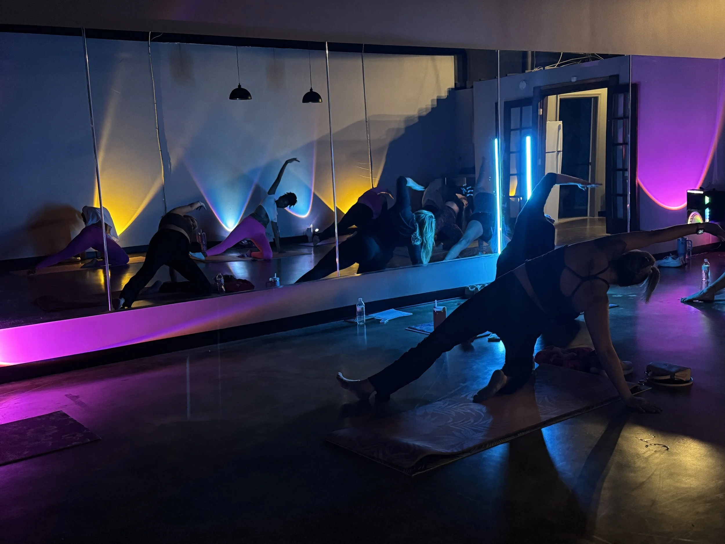 People practicing yoga or stretching on mats in a dimly lit studio with colorful neon and LED lighting, and a large mirror reflecting their poses.